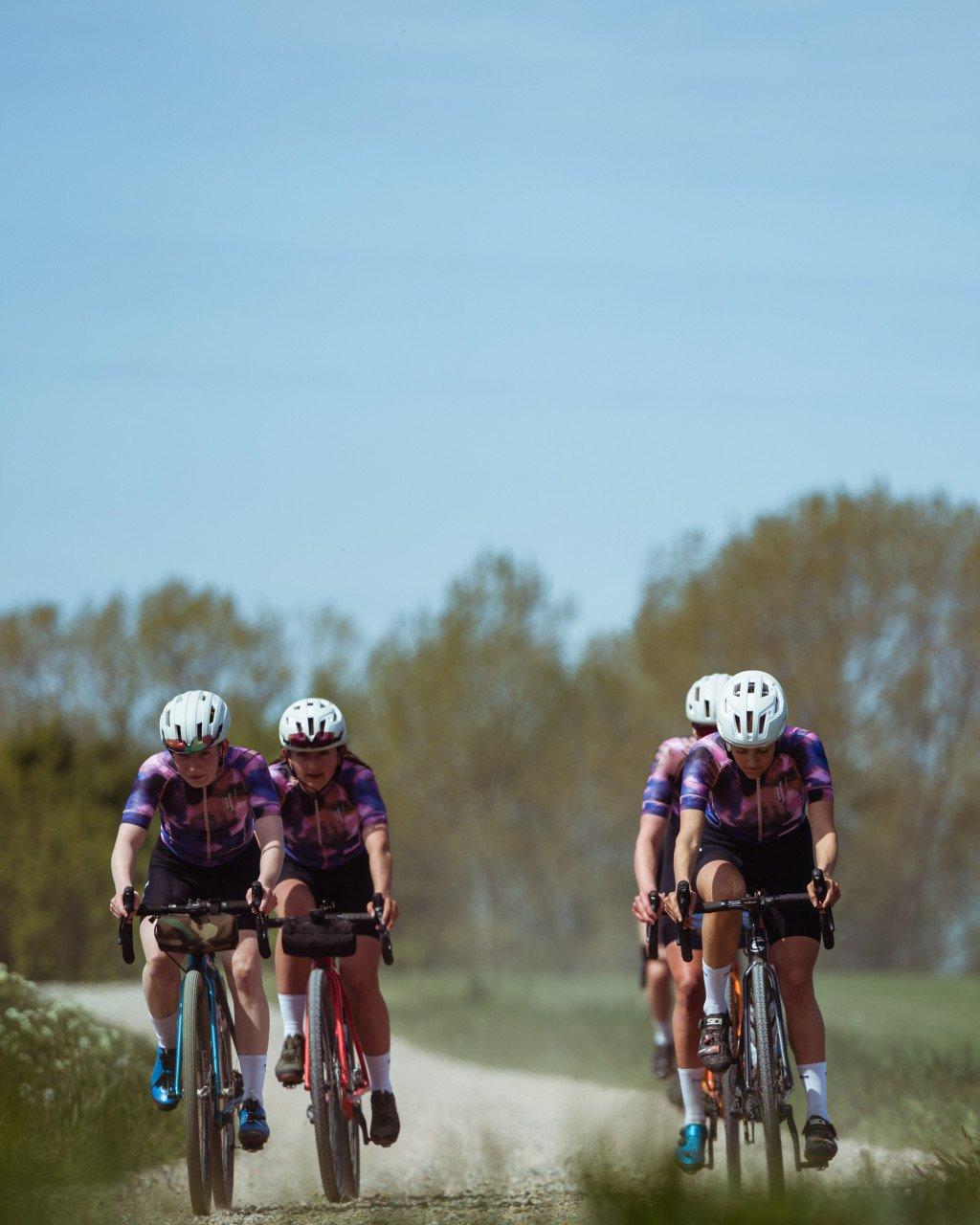 Cyclists in purple tie-dye jerseys riding time trial bikes on gravel road