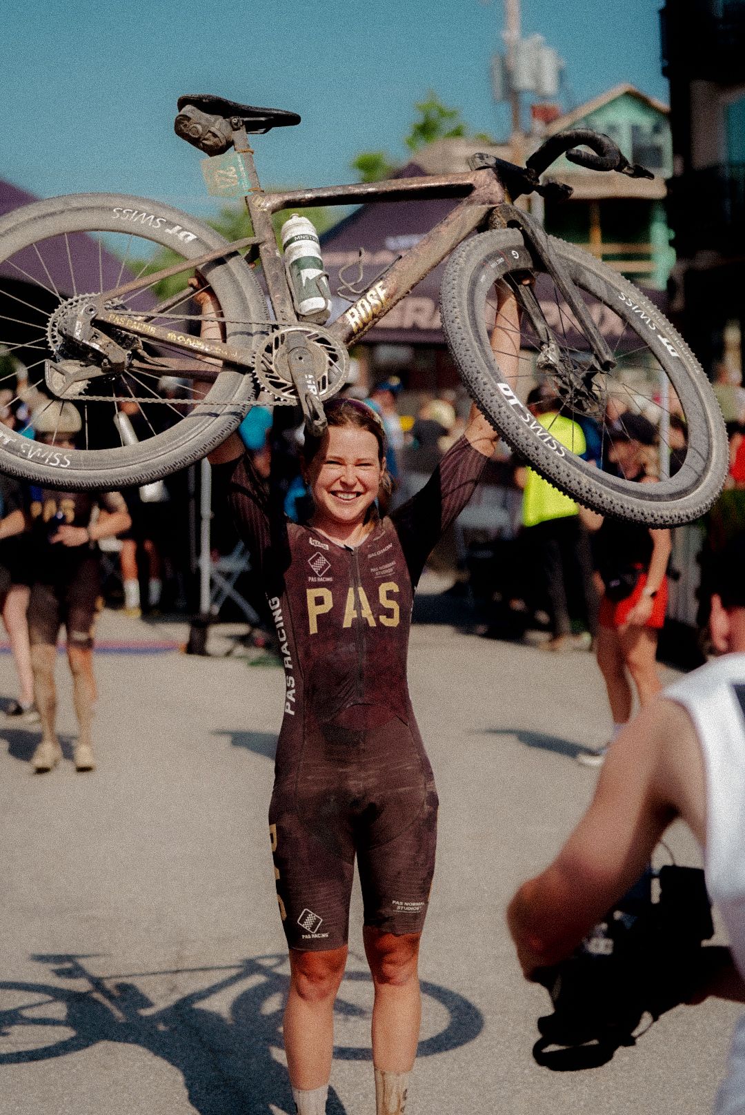 Model wearing Women's PAS collection brown skinsuit with chest branding, celebrating with bike raised overhead at gravel race finish