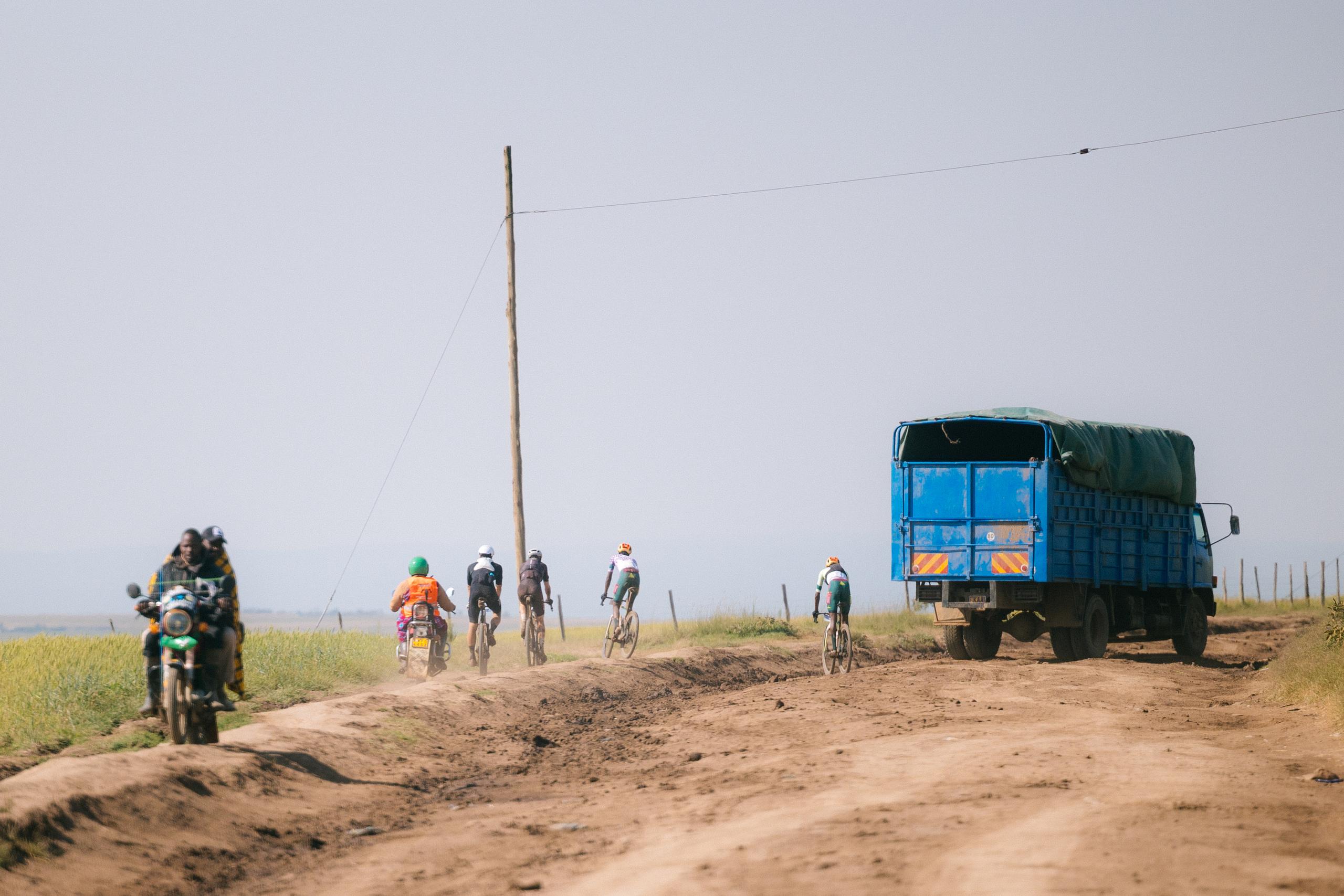 Cyclists racing on dirt road past blue truck in rural landscape