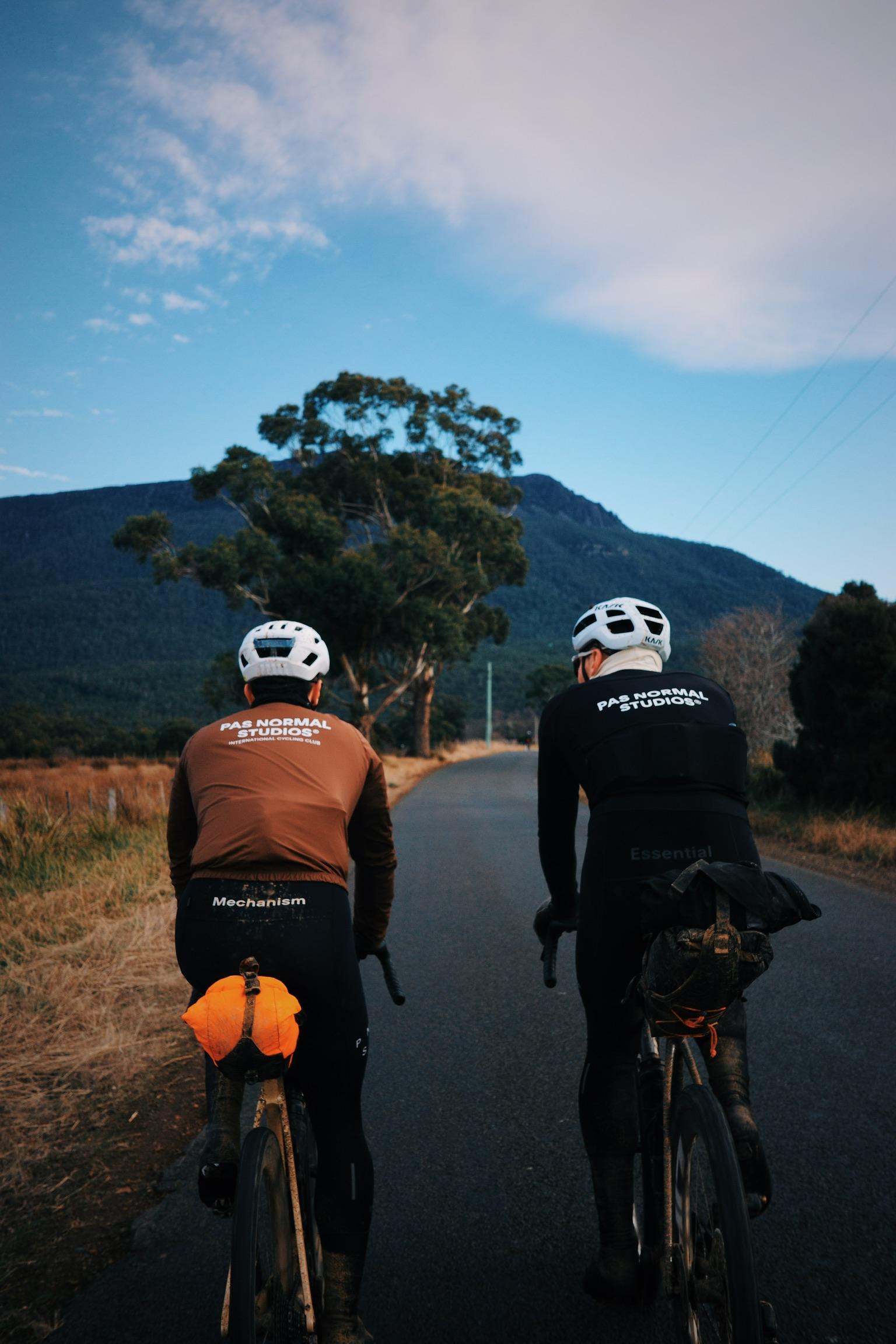 Two cyclists on road with brown and black long-sleeve jerseys with logo branding, mountain landscape background