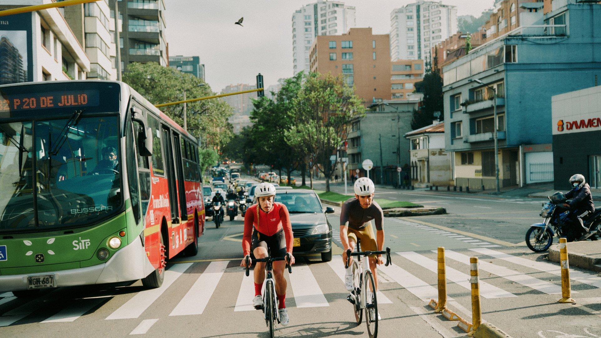 Two cyclists in time trial position crossing urban street intersection with bus and city buildings
