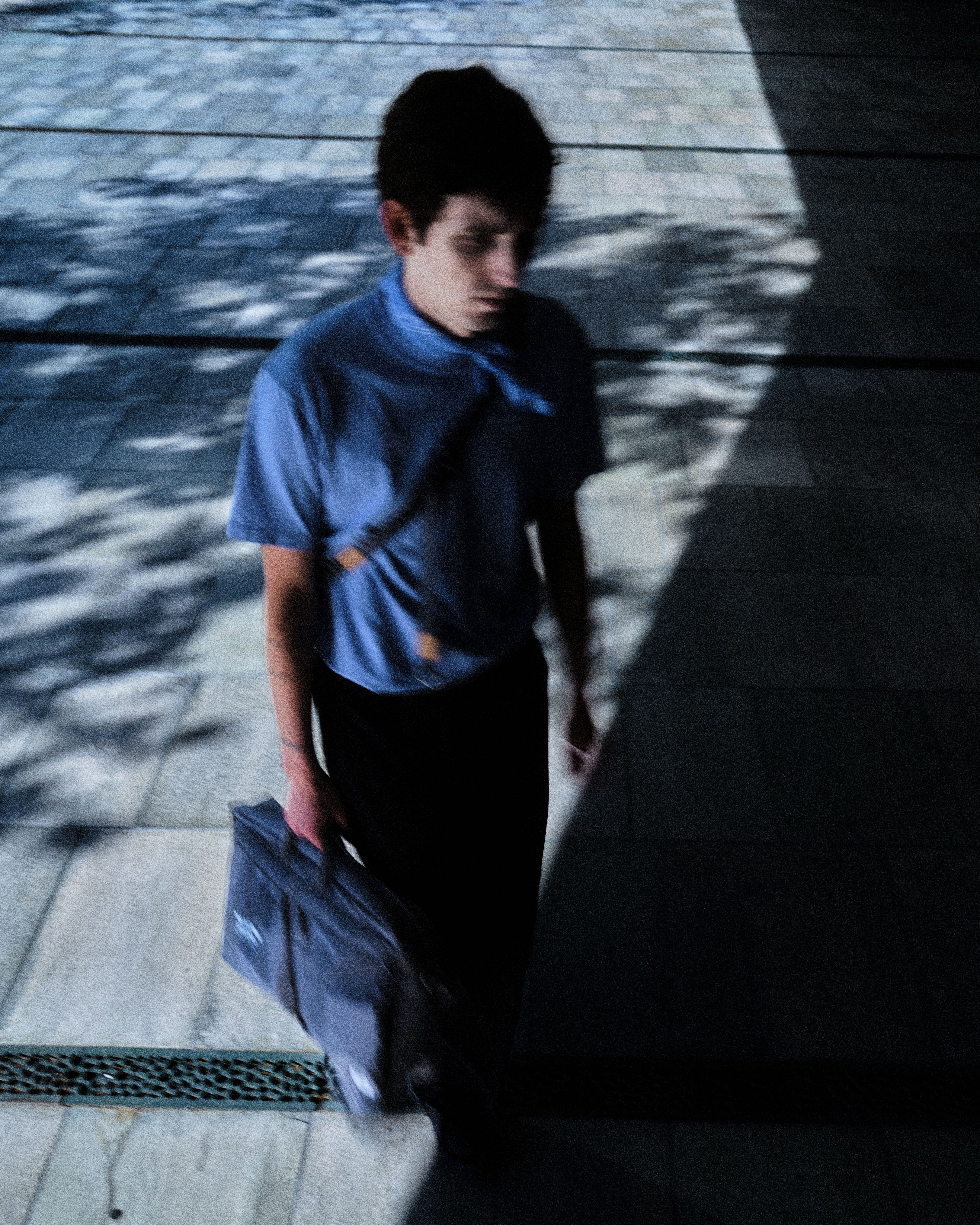 Model wearing blue t-shirt and black shorts carrying blue Porter-Yoshida bag in shadowed urban setting
