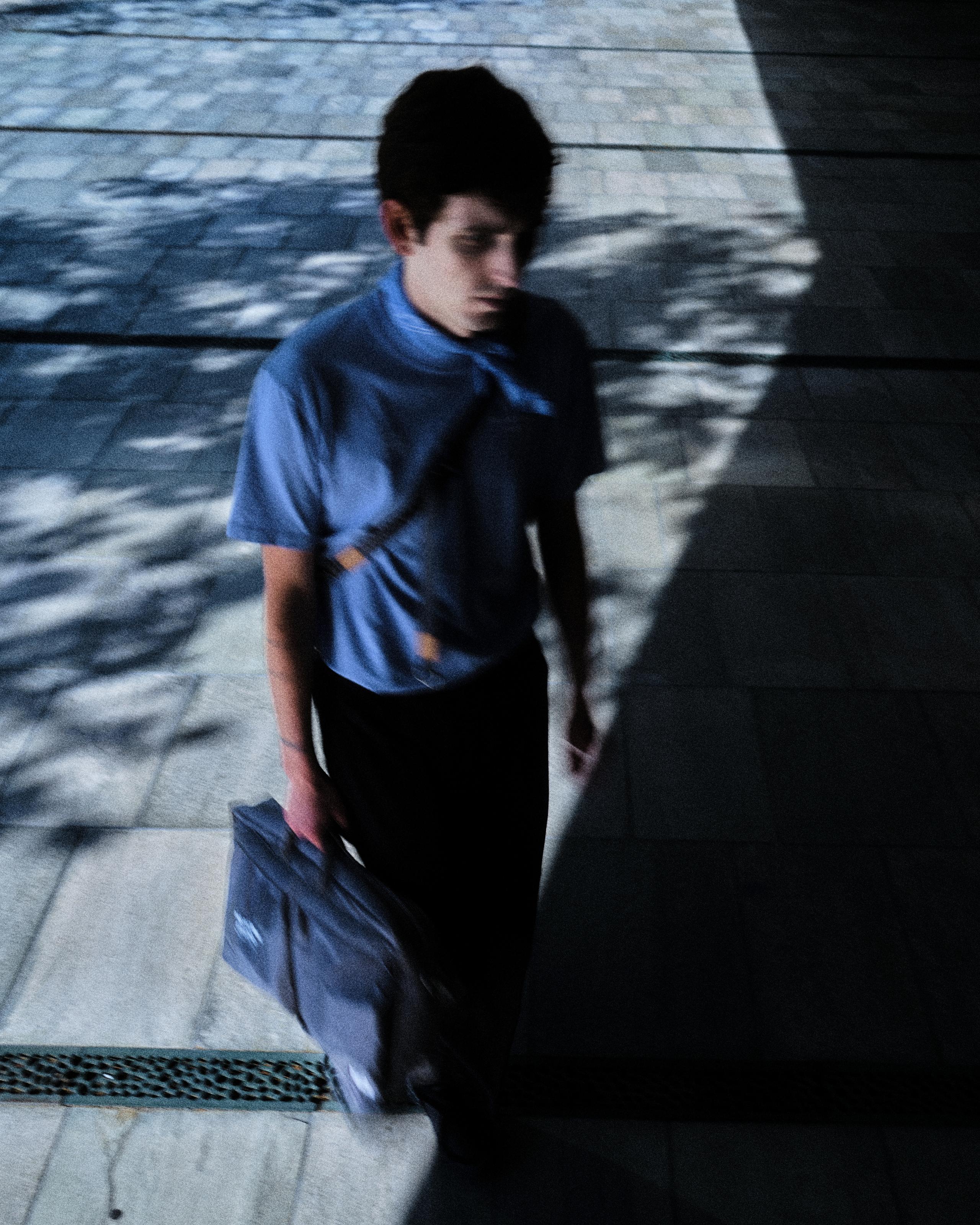 Model wearing blue t-shirt and black shorts carrying blue Porter-Yoshida bag in shadowed urban setting
