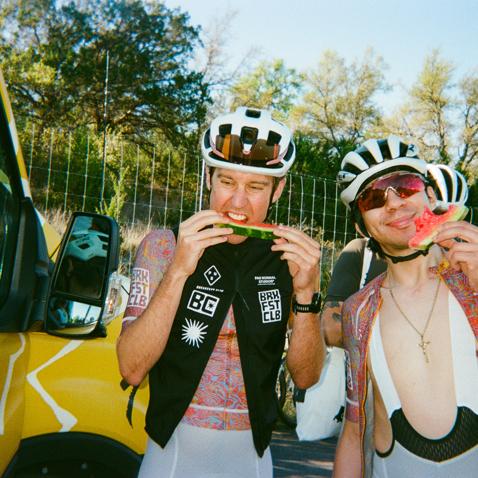 Two cyclists in white helmets eating watermelon beside yellow van, one wearing black jersey with white graphics