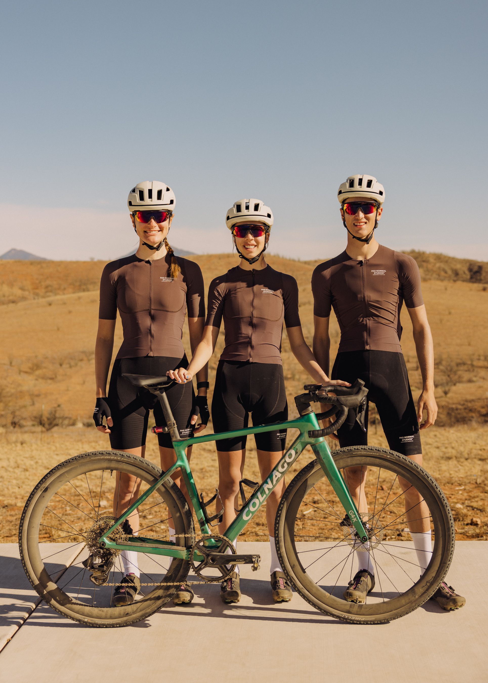 Three cyclists in brown jerseys and black shorts with green gravel bike in desert landscape