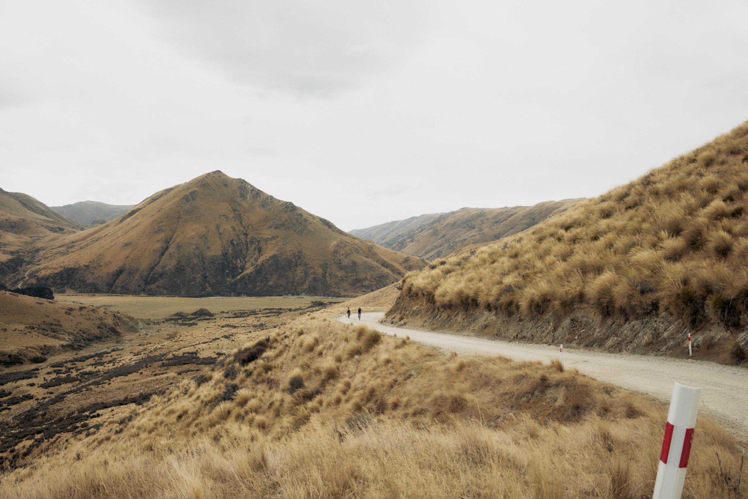 Two cyclists on winding mountain road through golden grassland hills under overcast sky