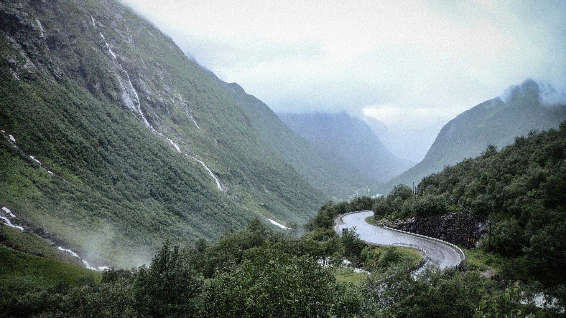 Winding mountain road through green Norwegian valley with misty peaks and waterfalls