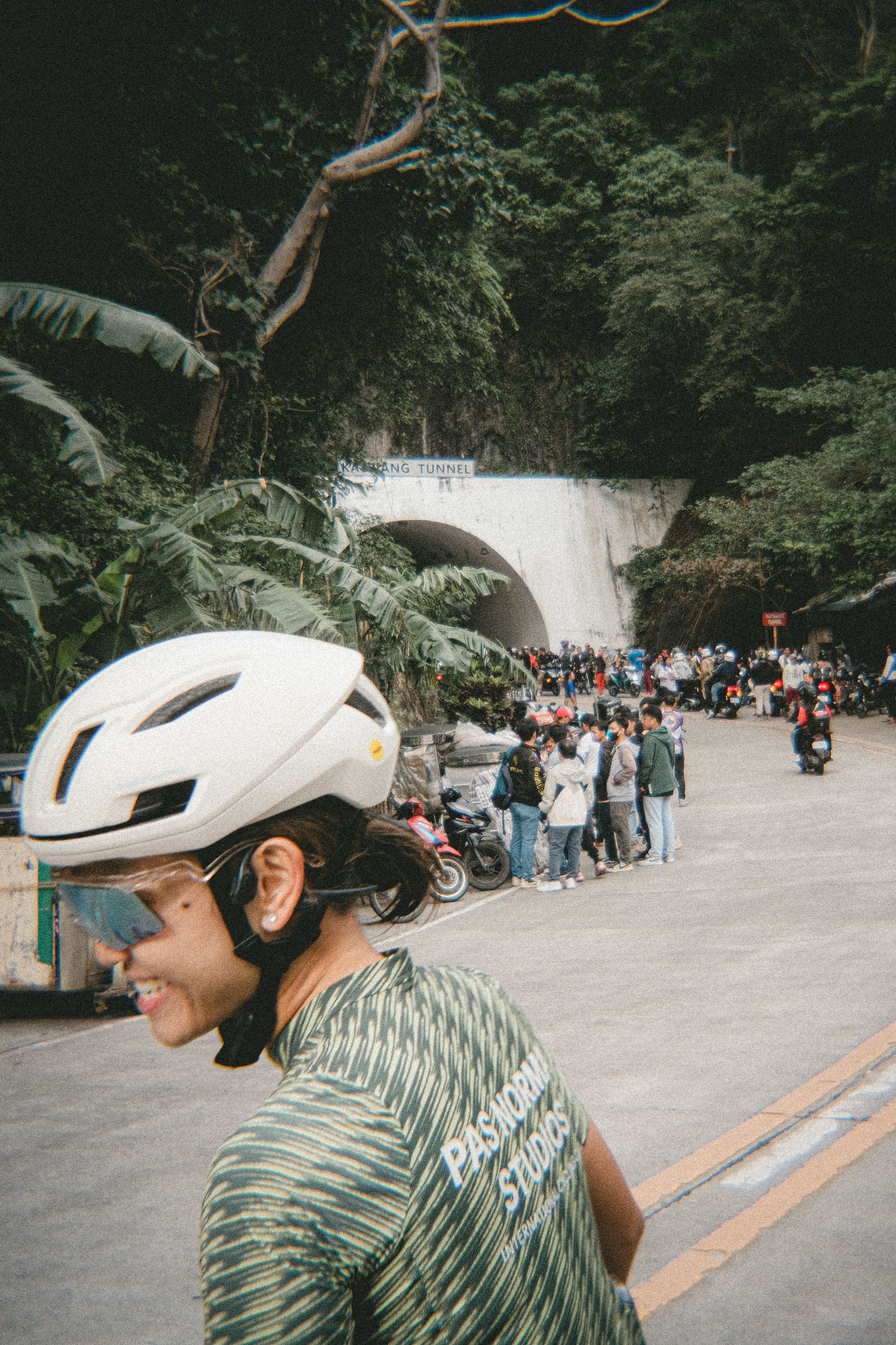 Model wearing green patterned jersey and white helmet cycling near tunnel entrance with crowd gathered