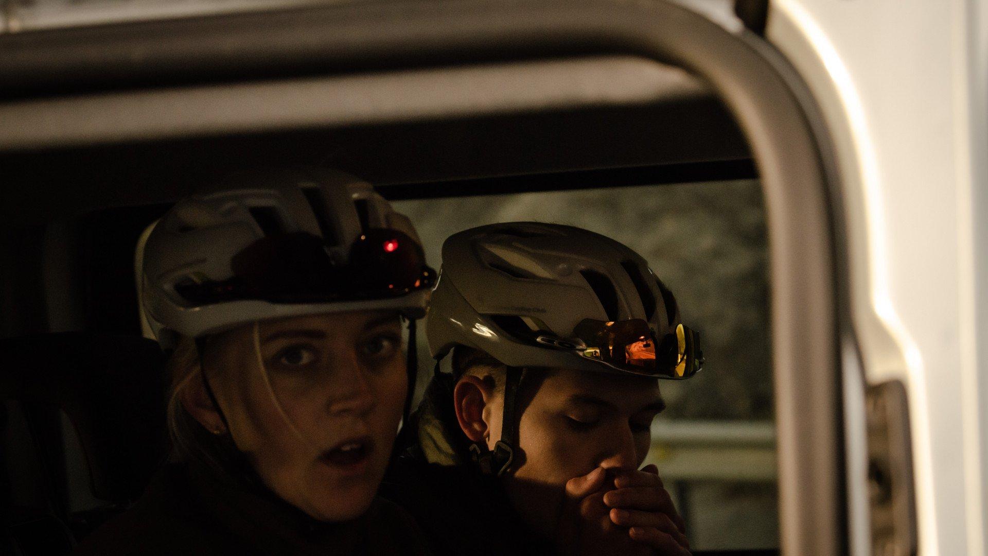 Two cyclists wearing aerodynamic helmets inside vehicle during golden hour lighting