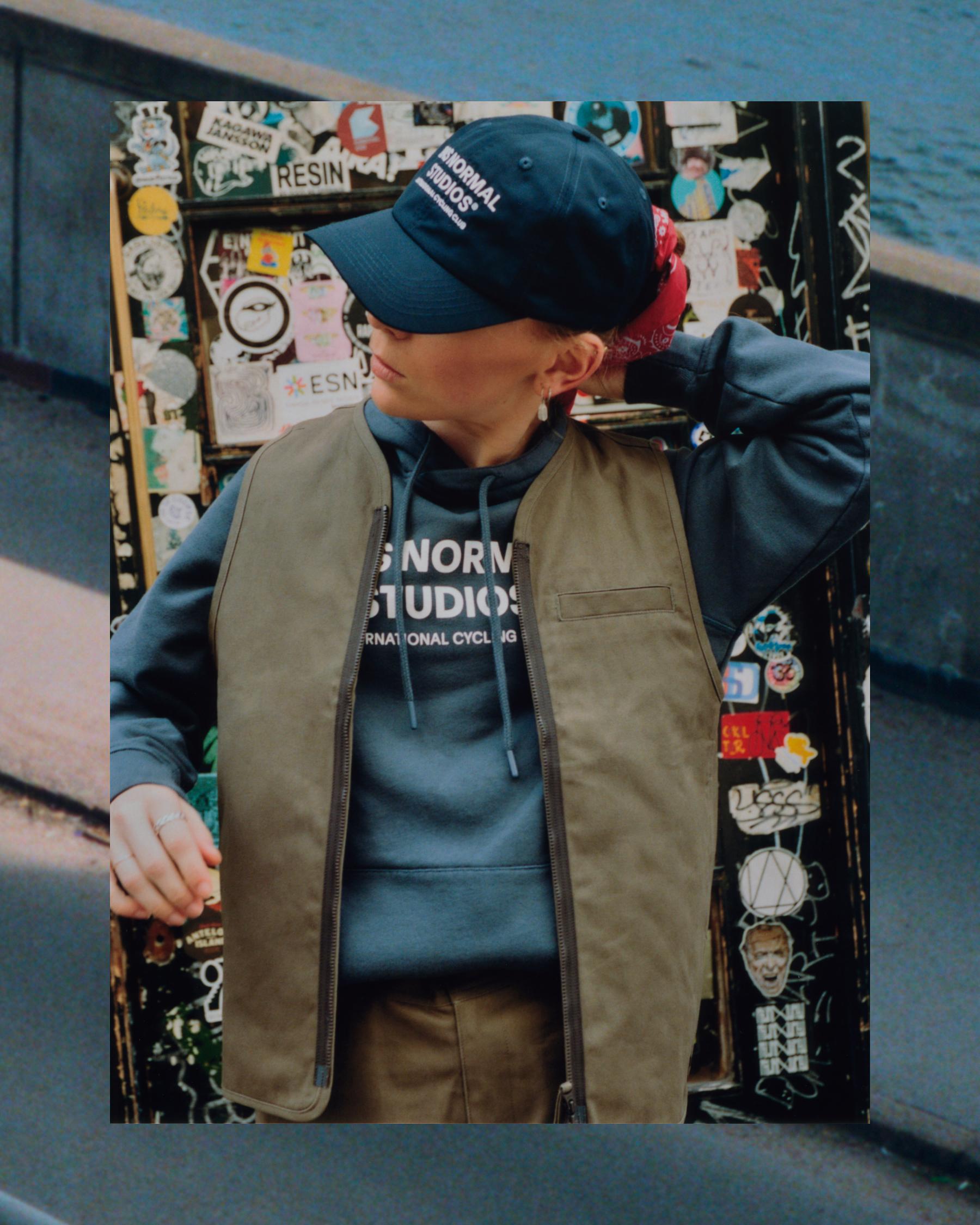 Model wearing navy cap with white lettering, blue hoodie, and tan zip vest in urban setting with sticker-covered wall