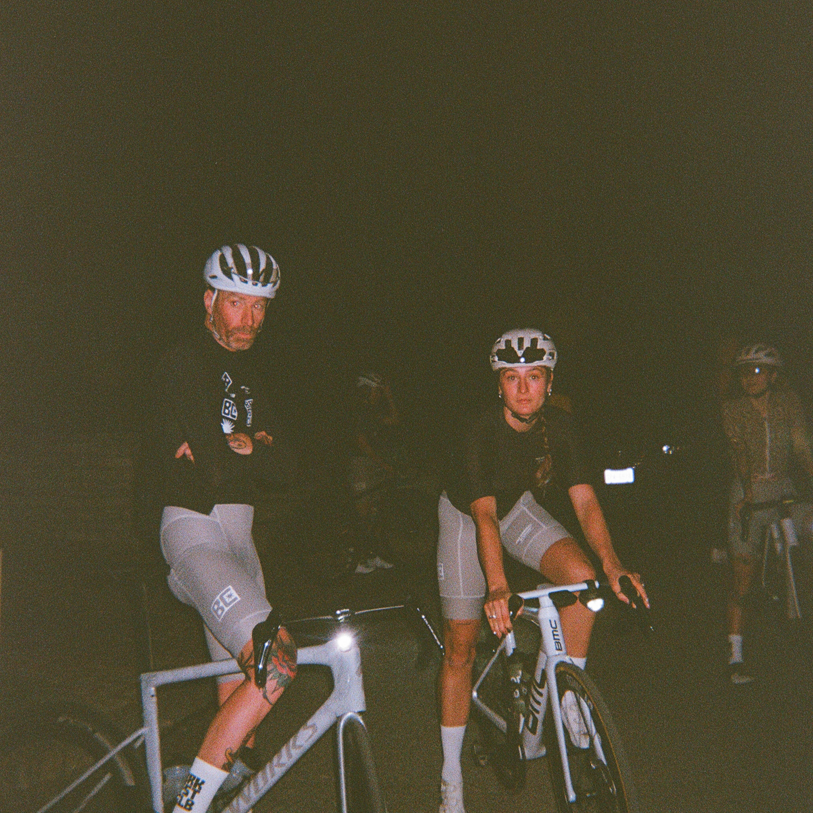 Two cyclists on indoor trainers wearing black jerseys and grey shorts during nighttime training session