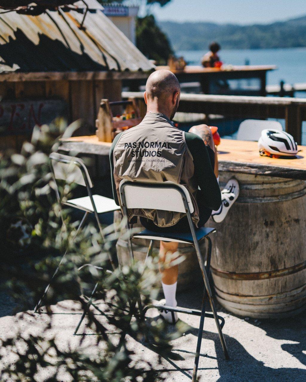 Model wearing beige cycling jacket with back branding, seated at lakeside cafe with cycling helmet nearby