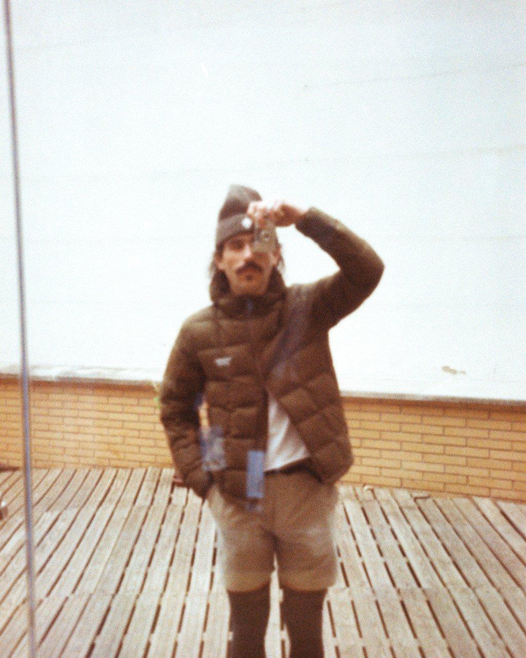 Model wearing brown down jacket with chest logo, standing on wooden deck with hand shielding eyes