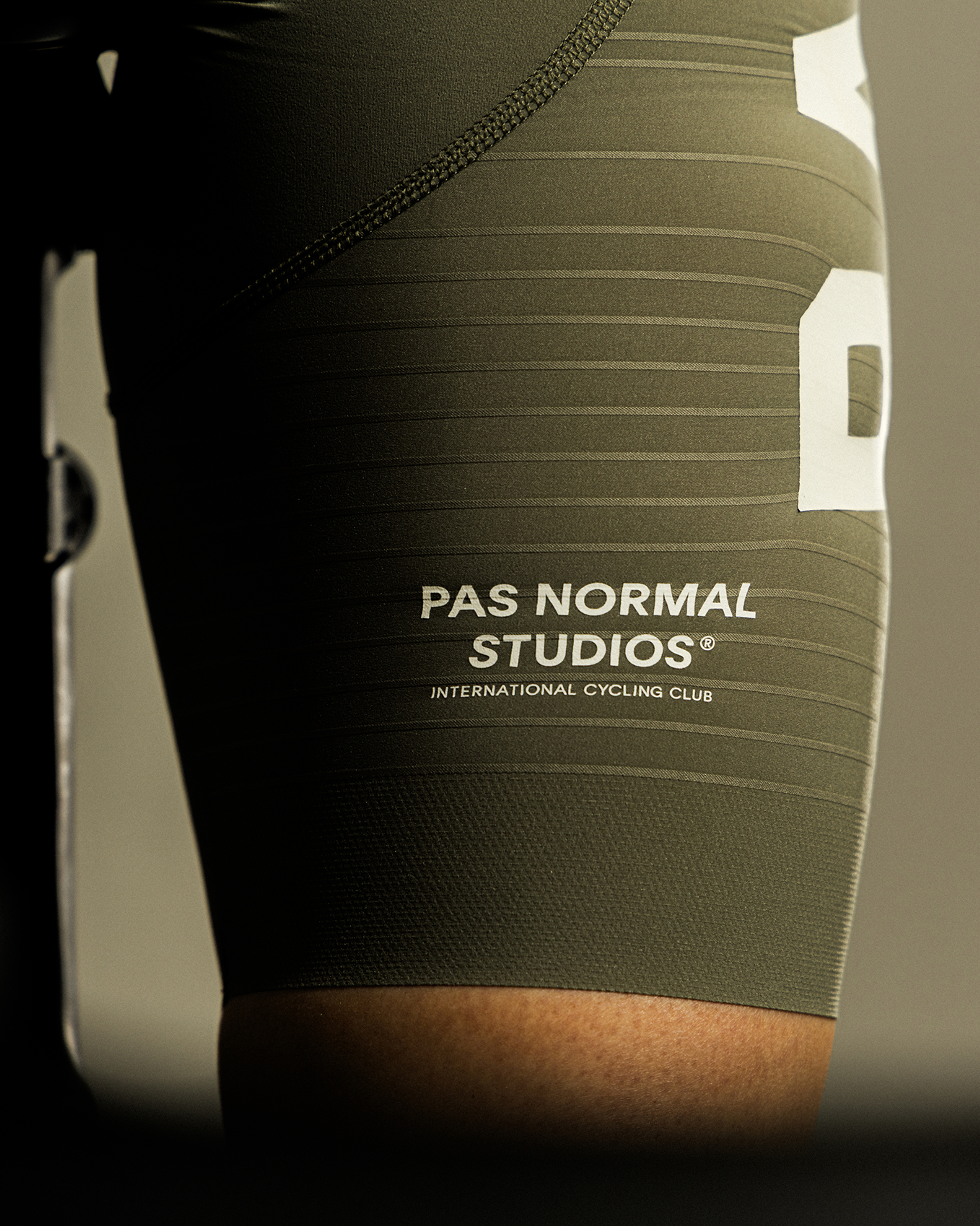 Model wearing olive green cycling bib shorts with horizontal stripe texture and white logo branding