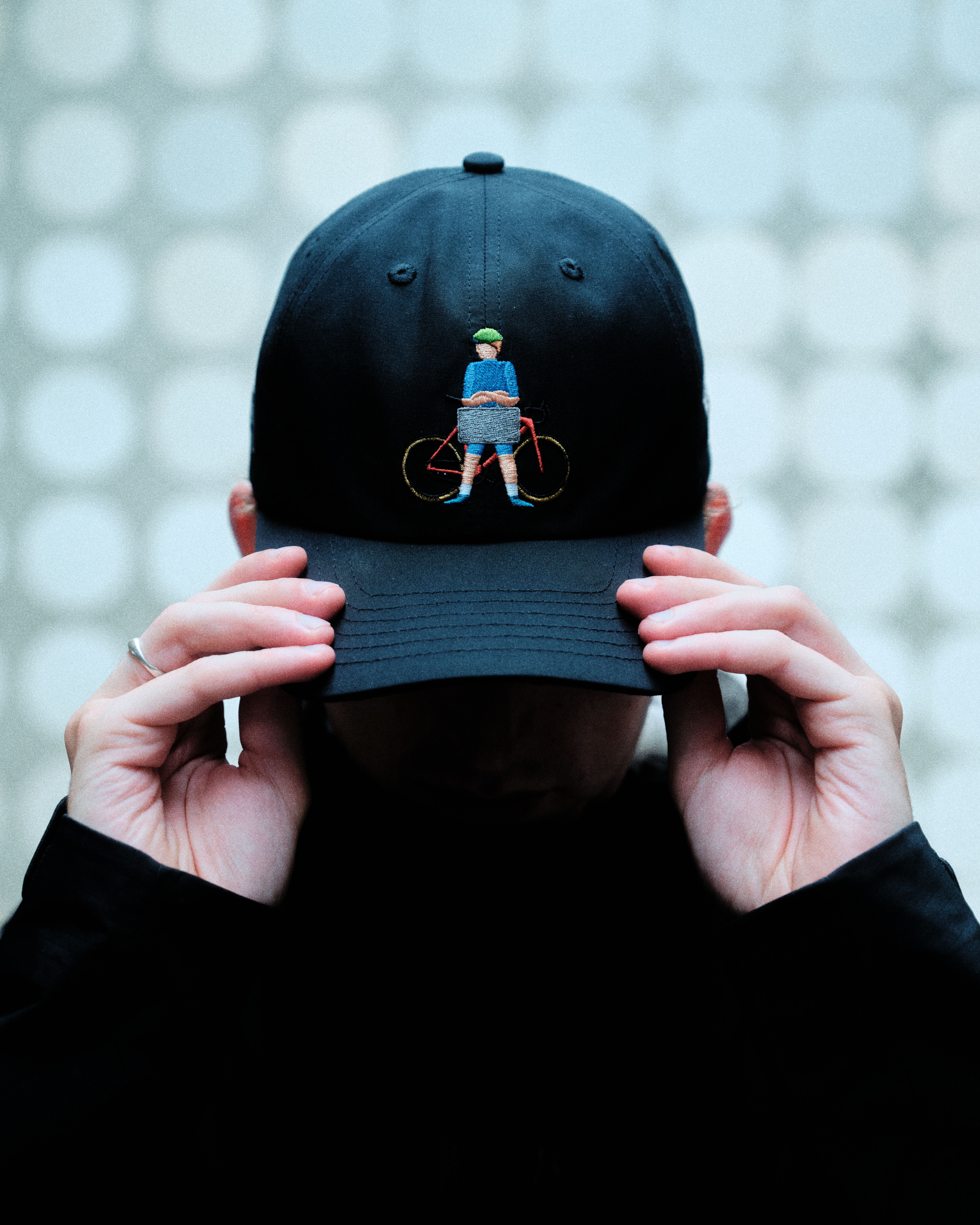 Model wearing black cap with embroidered cyclist figure on front panel