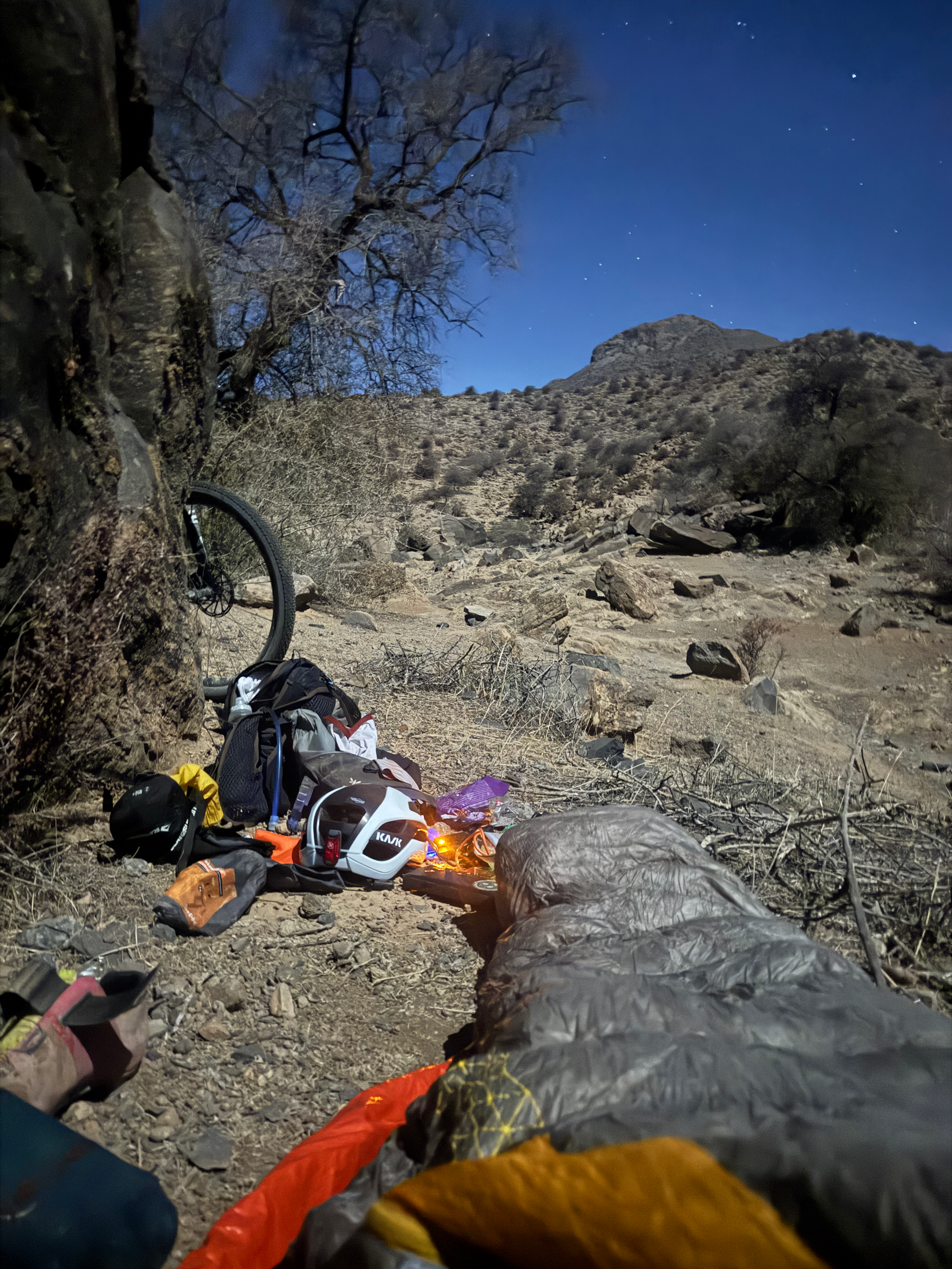 Bikepacking camp setup in desert landscape with scattered gear and bike wheel under starry sky