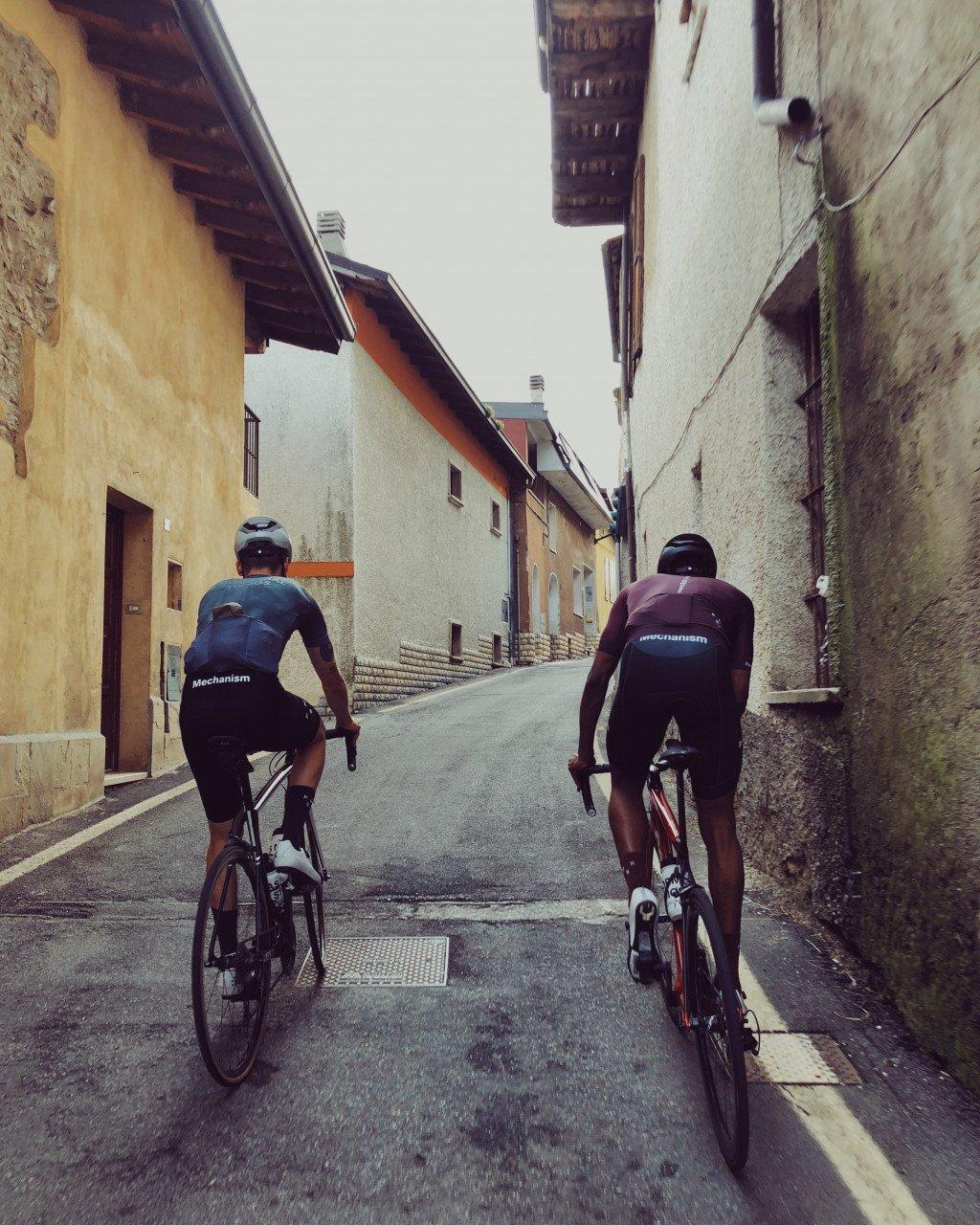 Two cyclists wearing Mechanism jerseys cycling through narrow European village street