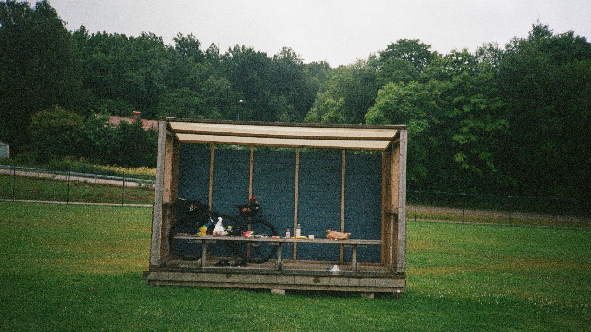 Cyclists resting at wooden shelter with bikes and supplies in green countryside setting