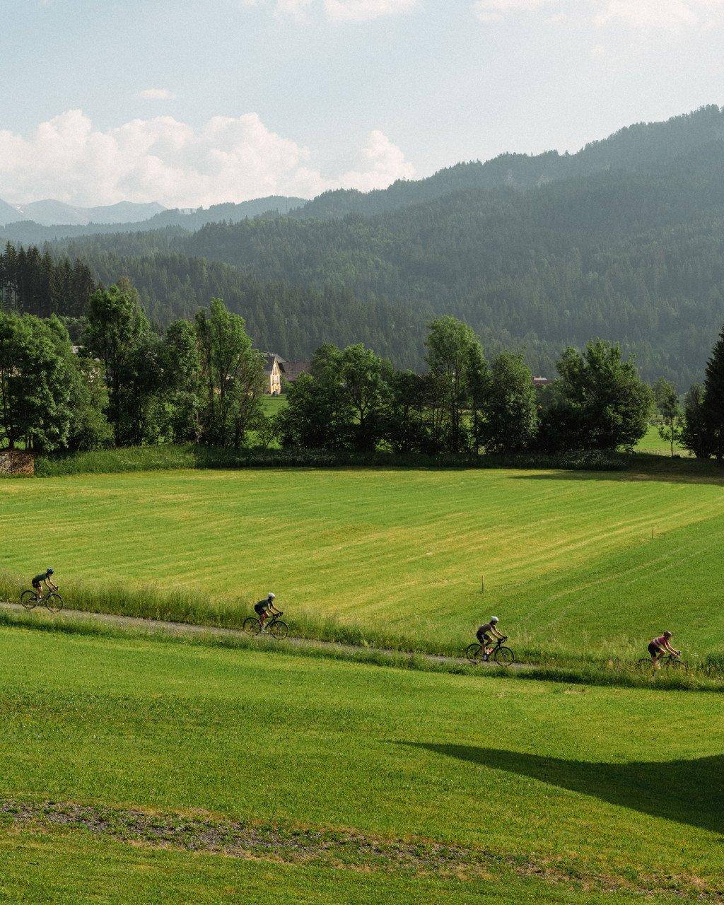 Four cyclists riding through green Austrian countryside with mountains and church in background