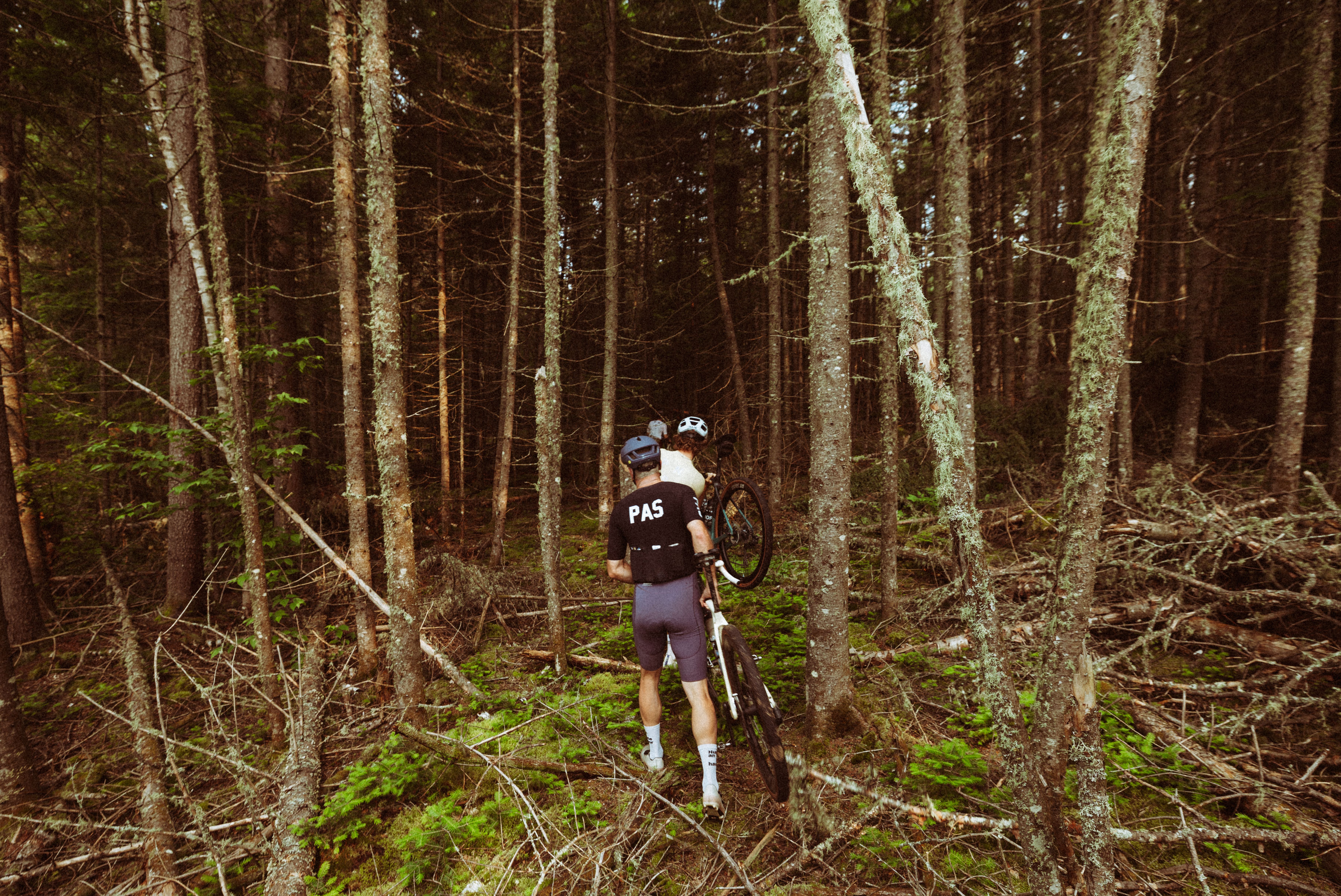 Cyclist carrying bike through forest wearing dark jersey and shorts with logo