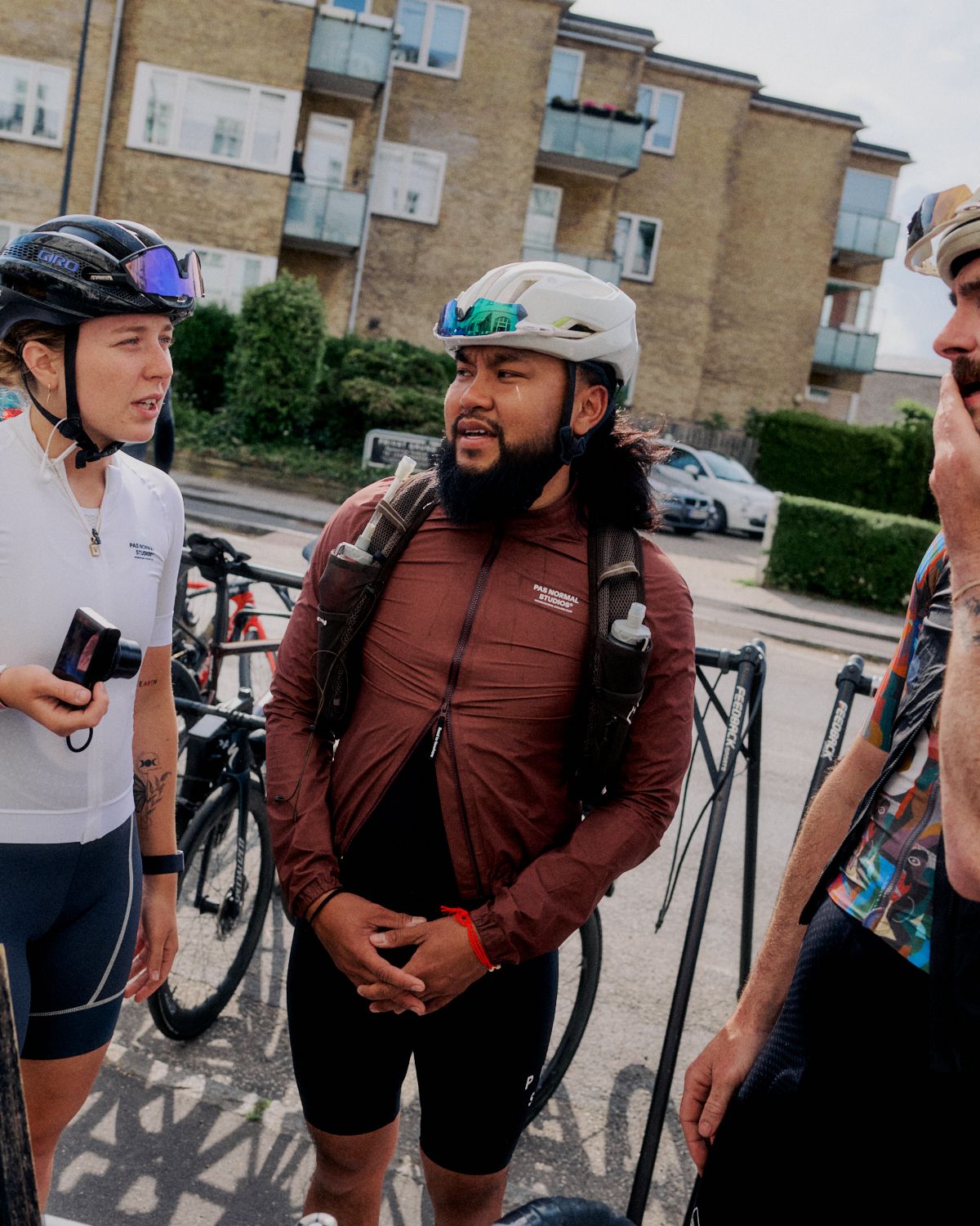 Three cyclists in urban setting wearing helmets and cycling apparel with bikes in background