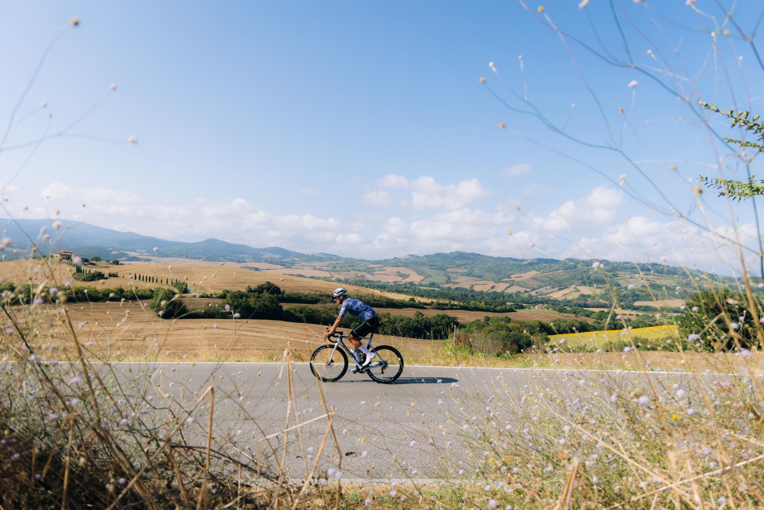 Model wearing blue cycling kit riding through rolling countryside landscape in Tuscany