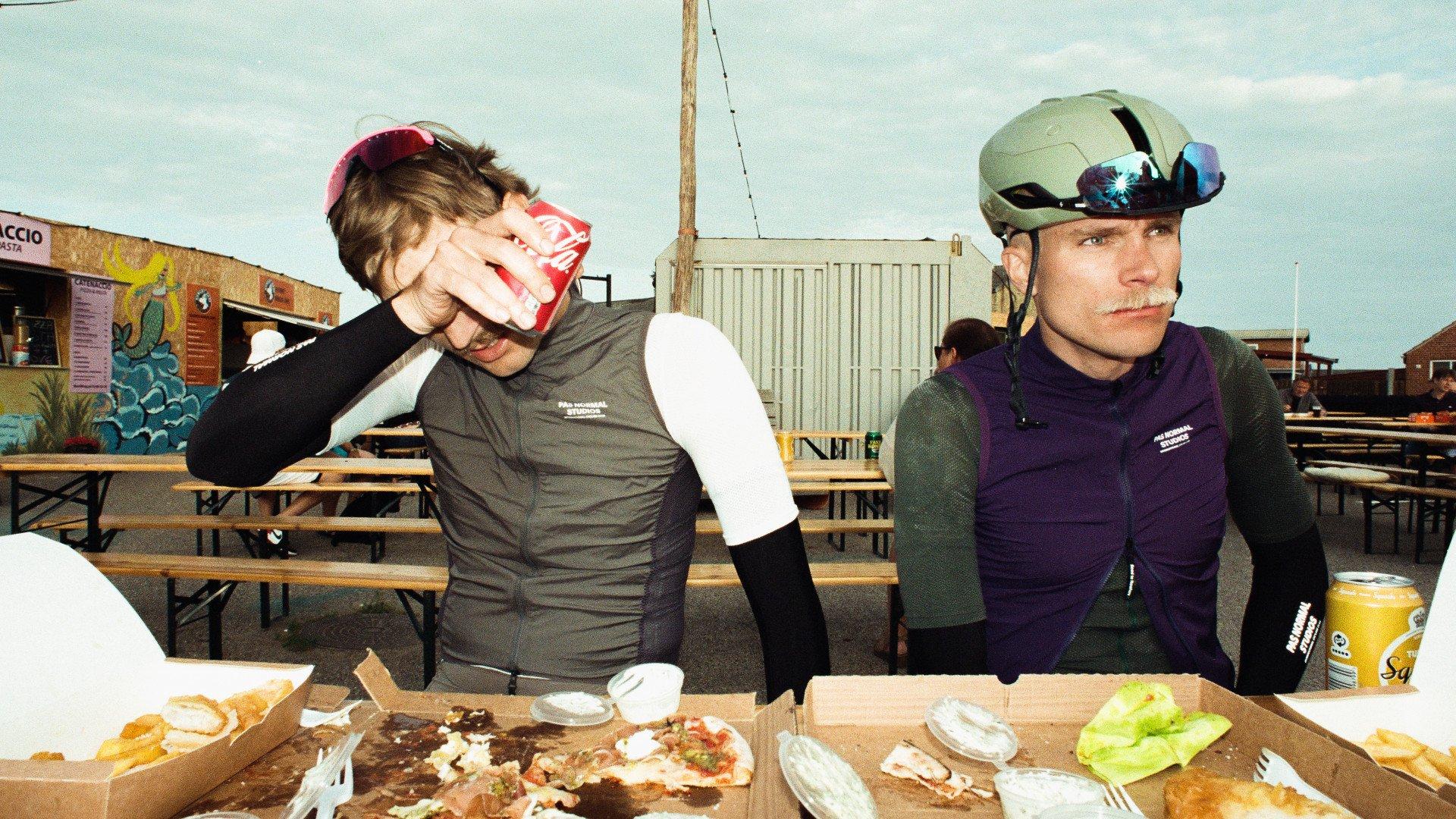 Two cyclists wearing cycling apparel eating at outdoor table with pizza and drinks