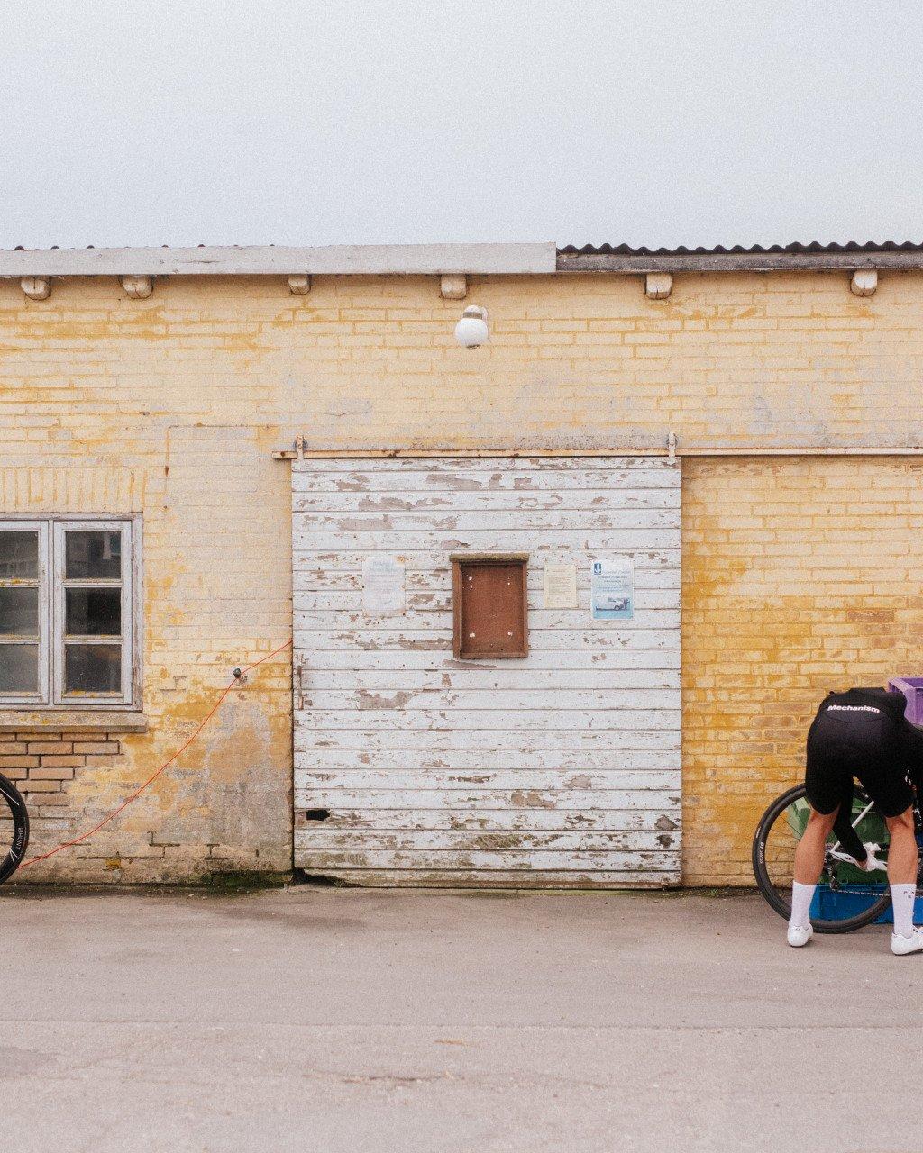 Cyclist in green jersey and black cap leaning against bike by weathered yellow brick building