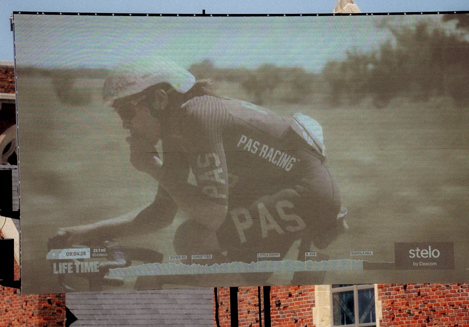 Large outdoor screen displaying cyclist in PAS Racing jersey during gravel race broadcast