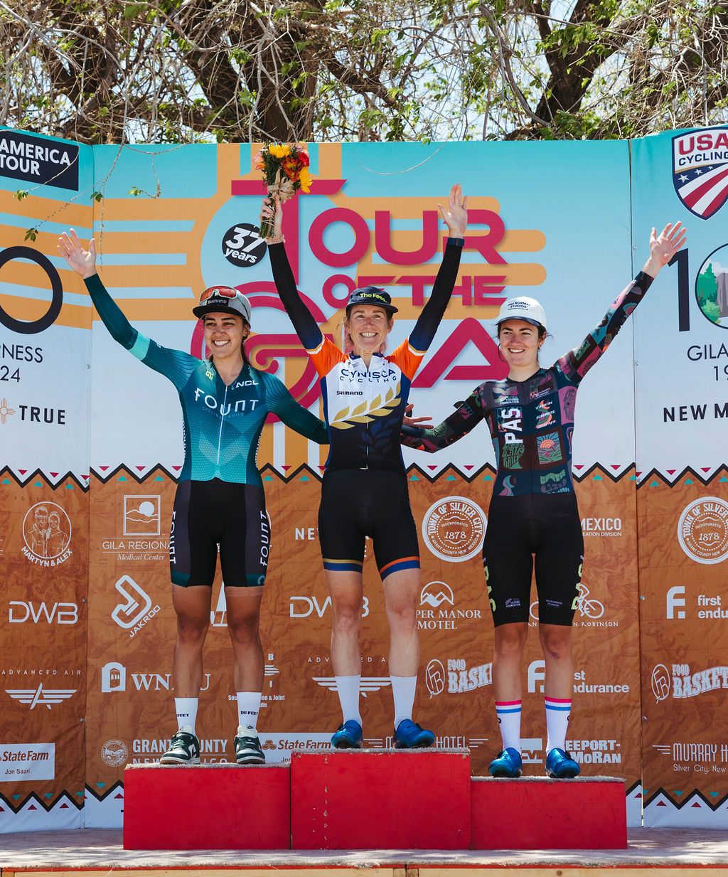 Three female cyclists on podium at Tour of the Gila, winner in center wearing orange and navy jersey with flower bouquet