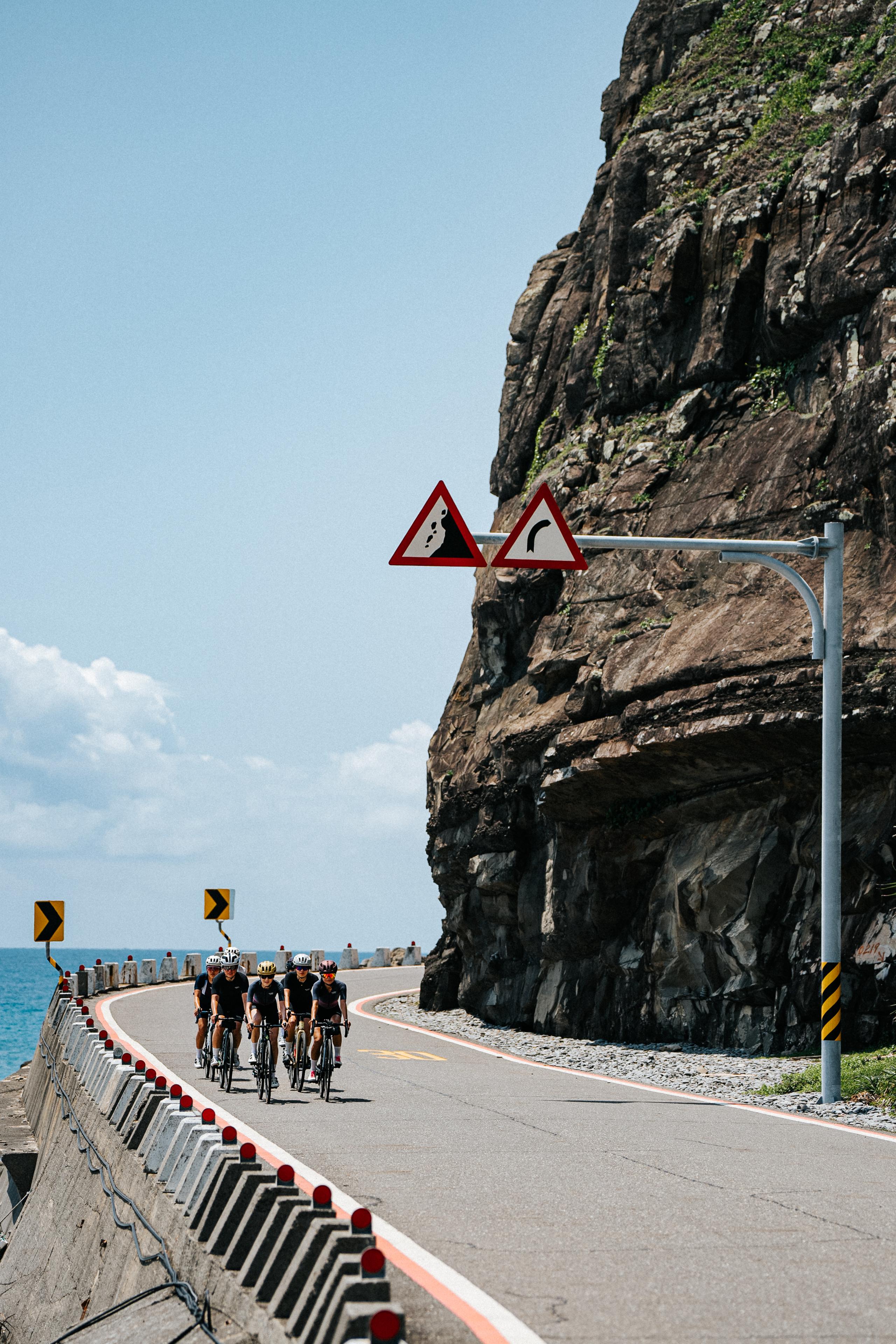 Group of cyclists riding on coastal mountain road with warning signs and ocean view