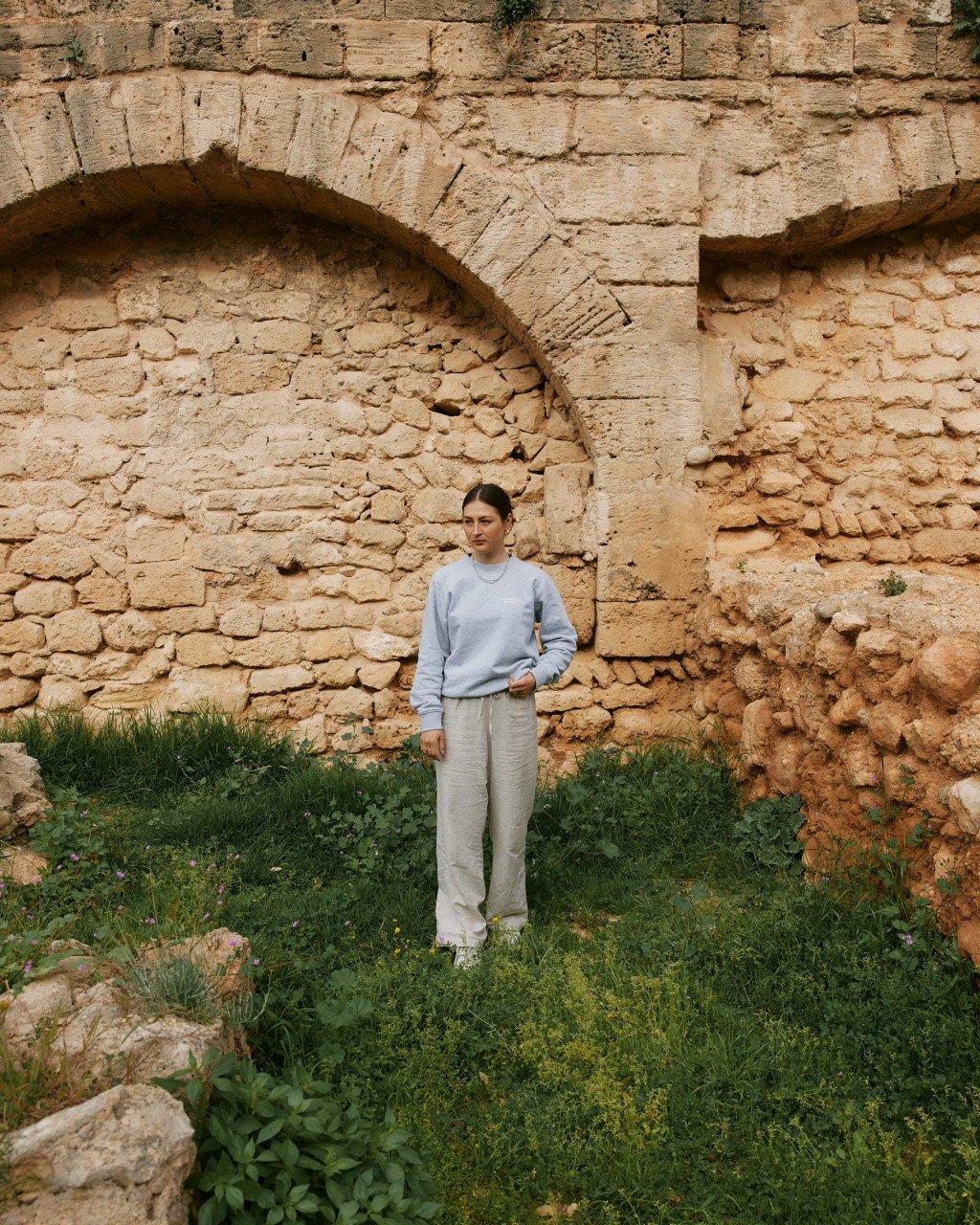 Model wearing light blue sweatshirt and cream trousers standing against ancient stone wall