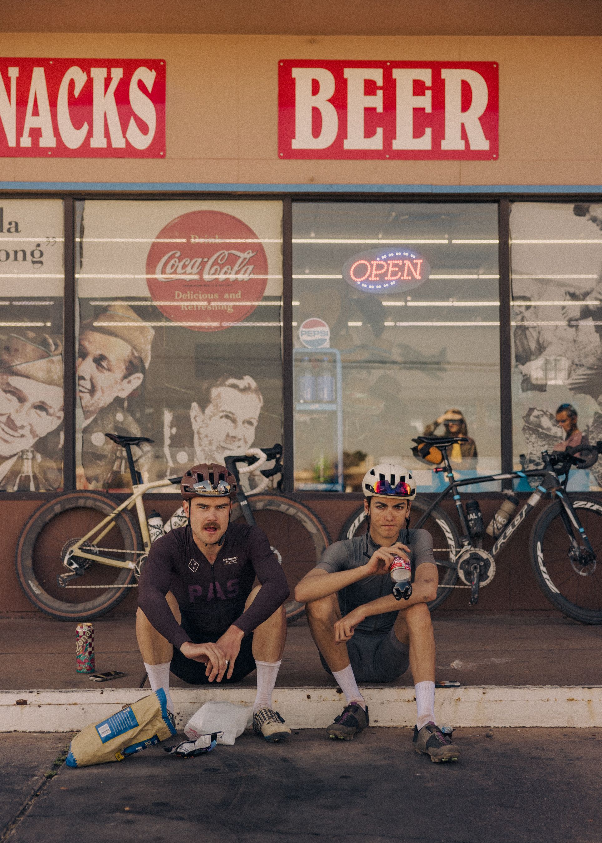 Two cyclists in helmets sitting outside convenience store with bikes, one wearing dark jersey with chest branding