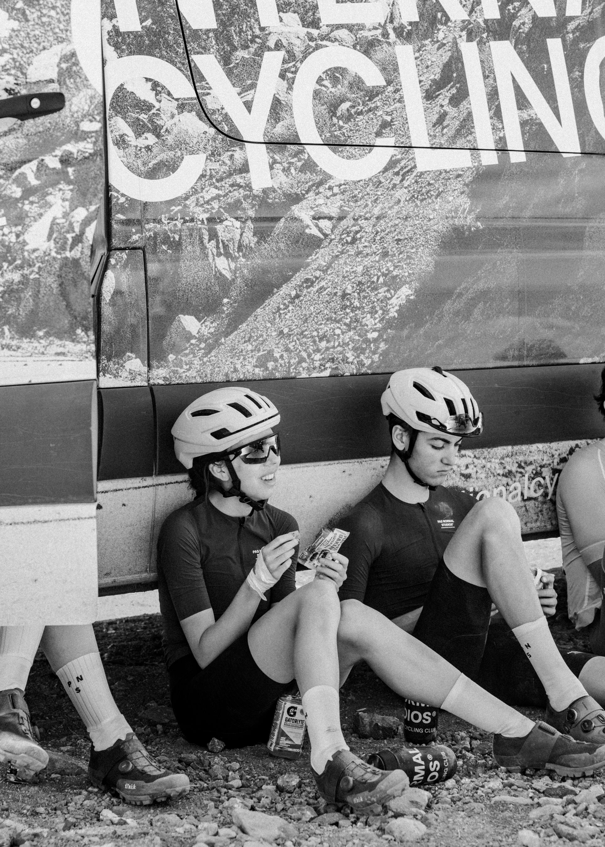 Two cyclists in dark jerseys and helmets sitting against car with cycling text, sharing post-ride moment