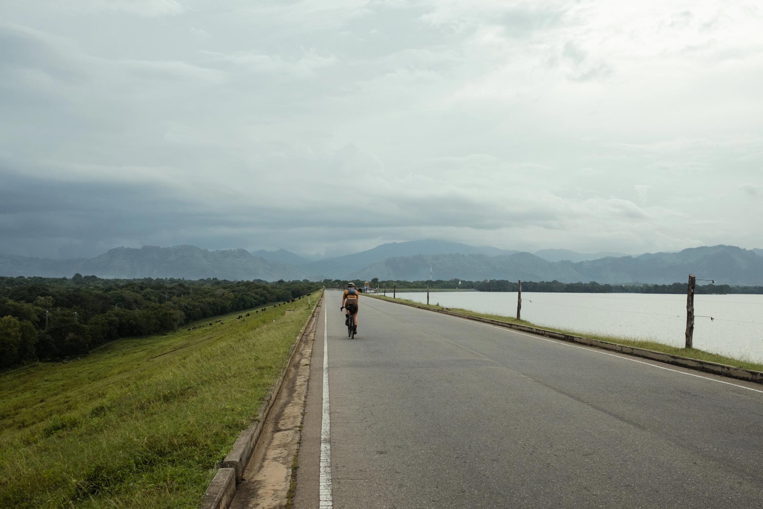 Cyclist riding on empty road between lake and mountains under overcast sky