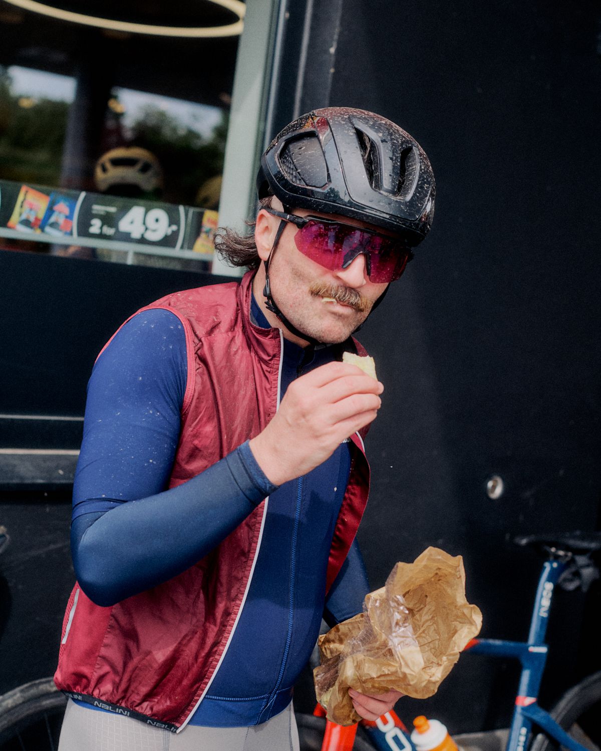 Model wearing blue long-sleeve jersey with burgundy vest, cycling helmet and sunglasses, eating at roadside stop