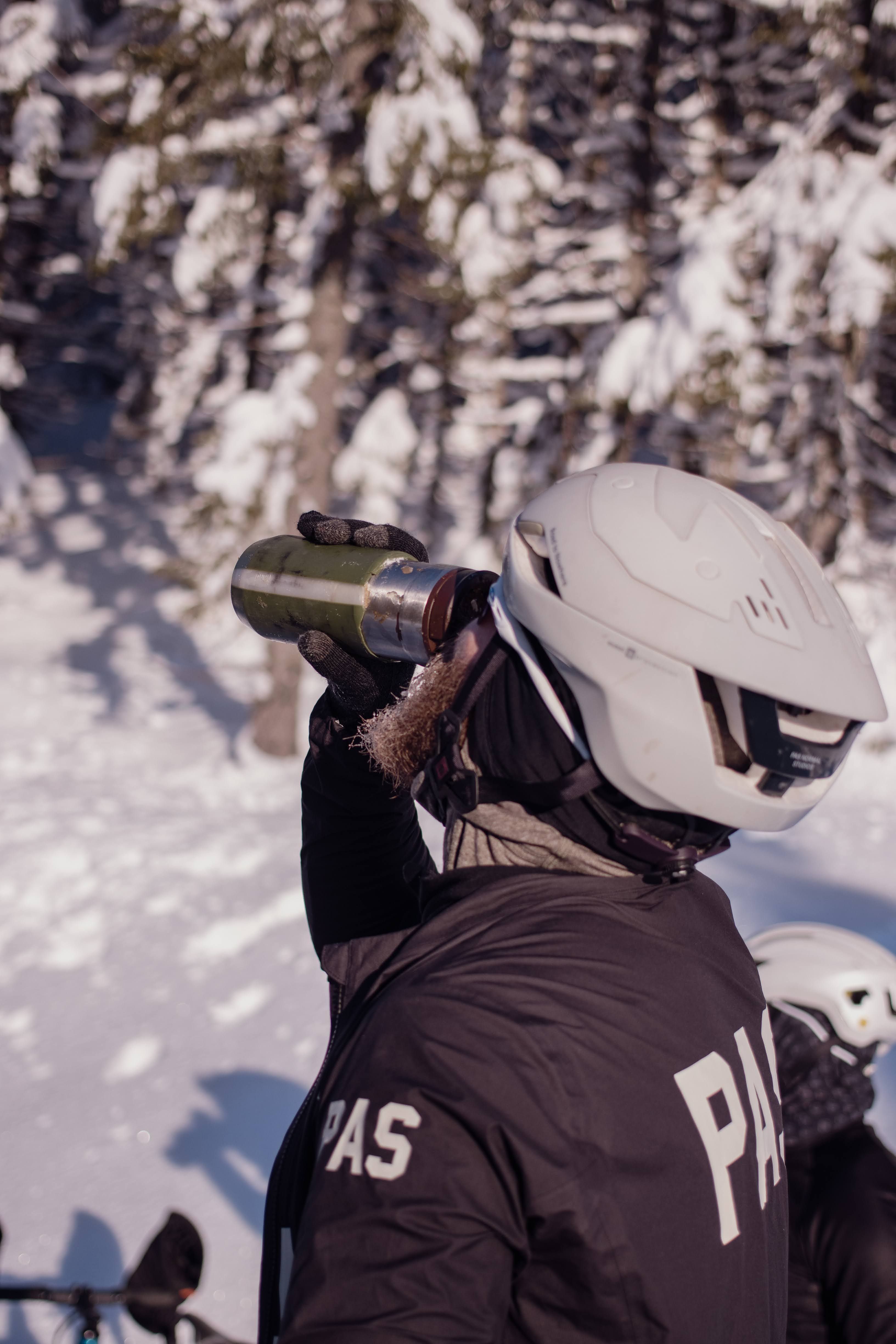 Model wearing brown thermal jacket with branding, holding bottle in snowy mountain setting
