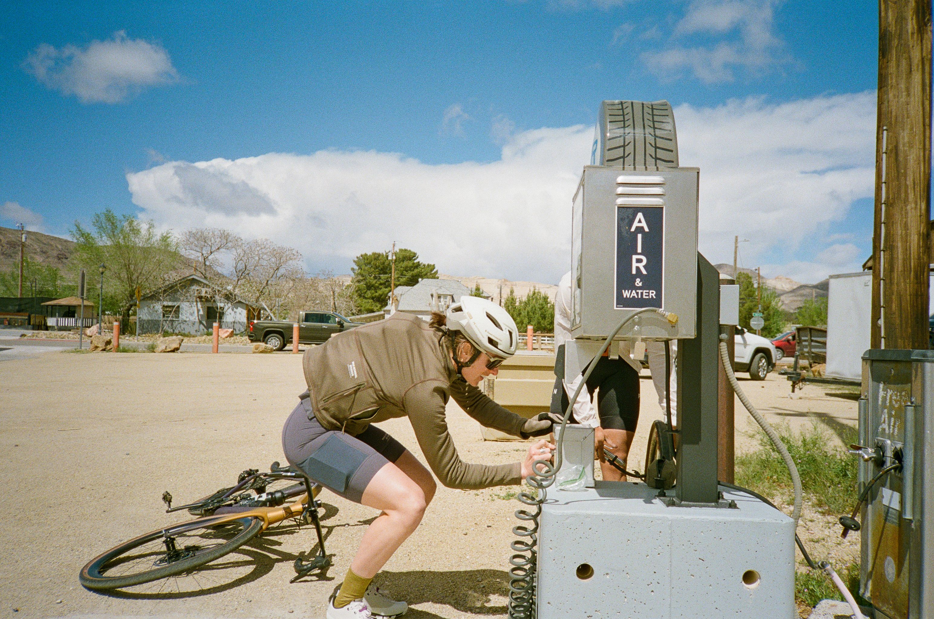 Cyclist in brown long-sleeve jersey and dark shorts filling water bottle at roadside station in desert town