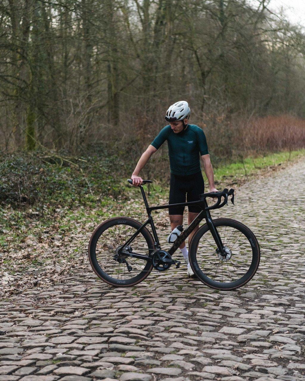 Model wearing dark green cycling jersey and black shorts with bike on cobblestone path