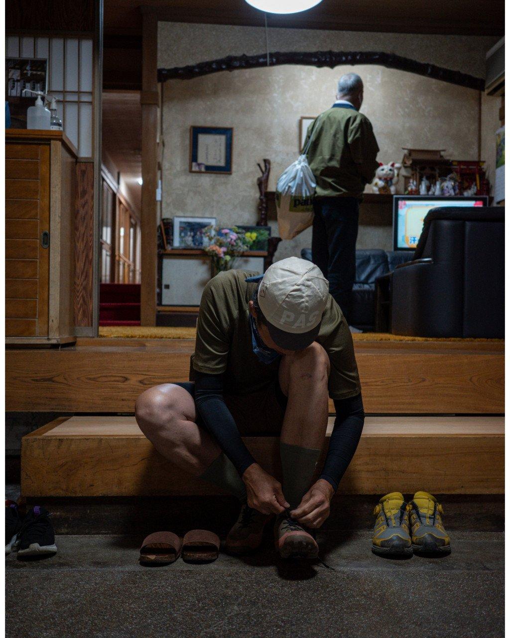 Model wearing green long-sleeve jersey and cap tying shoes in traditional Japanese interior