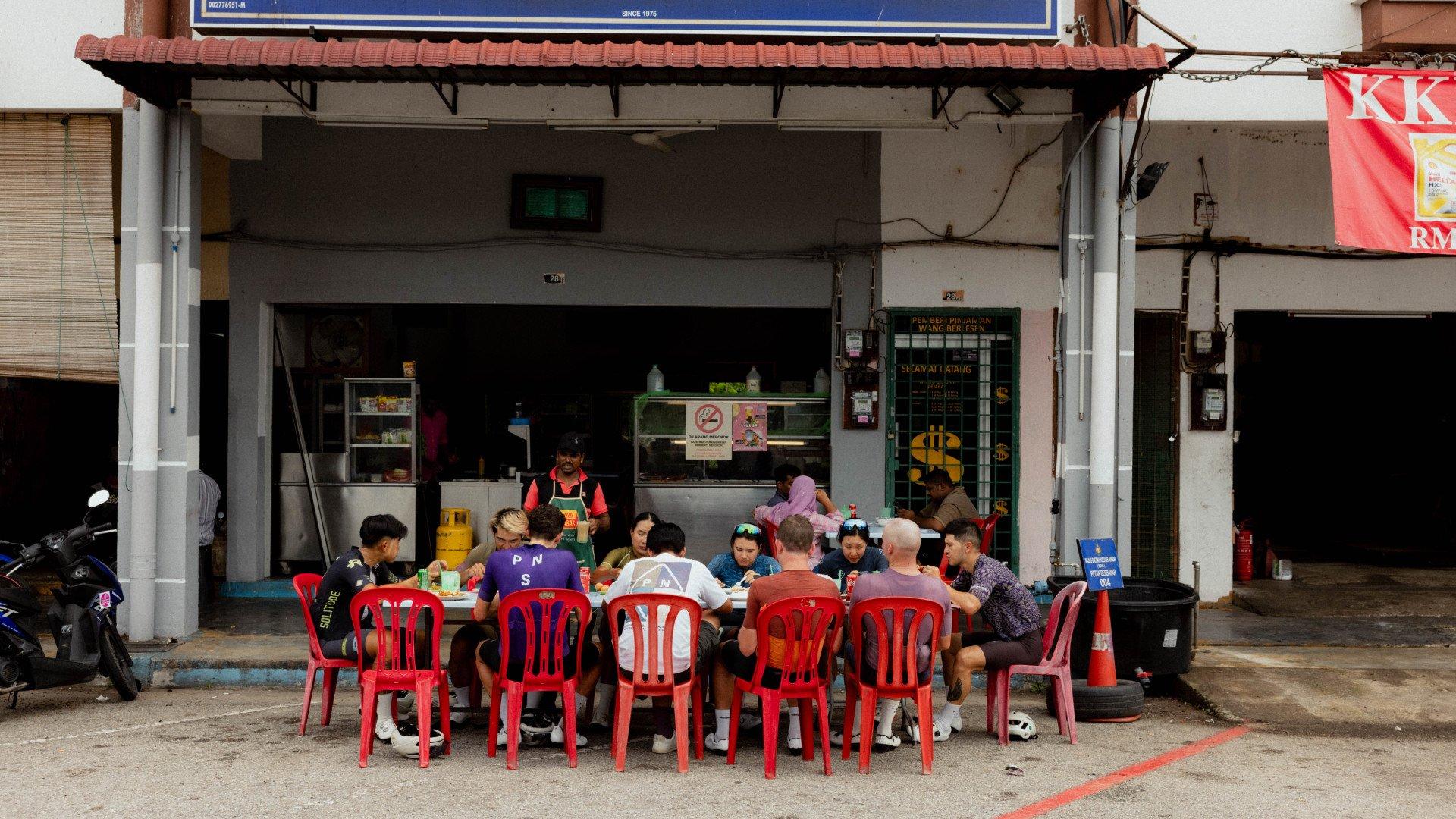Cyclists dining at local food stall with red plastic chairs in Singapore street setting