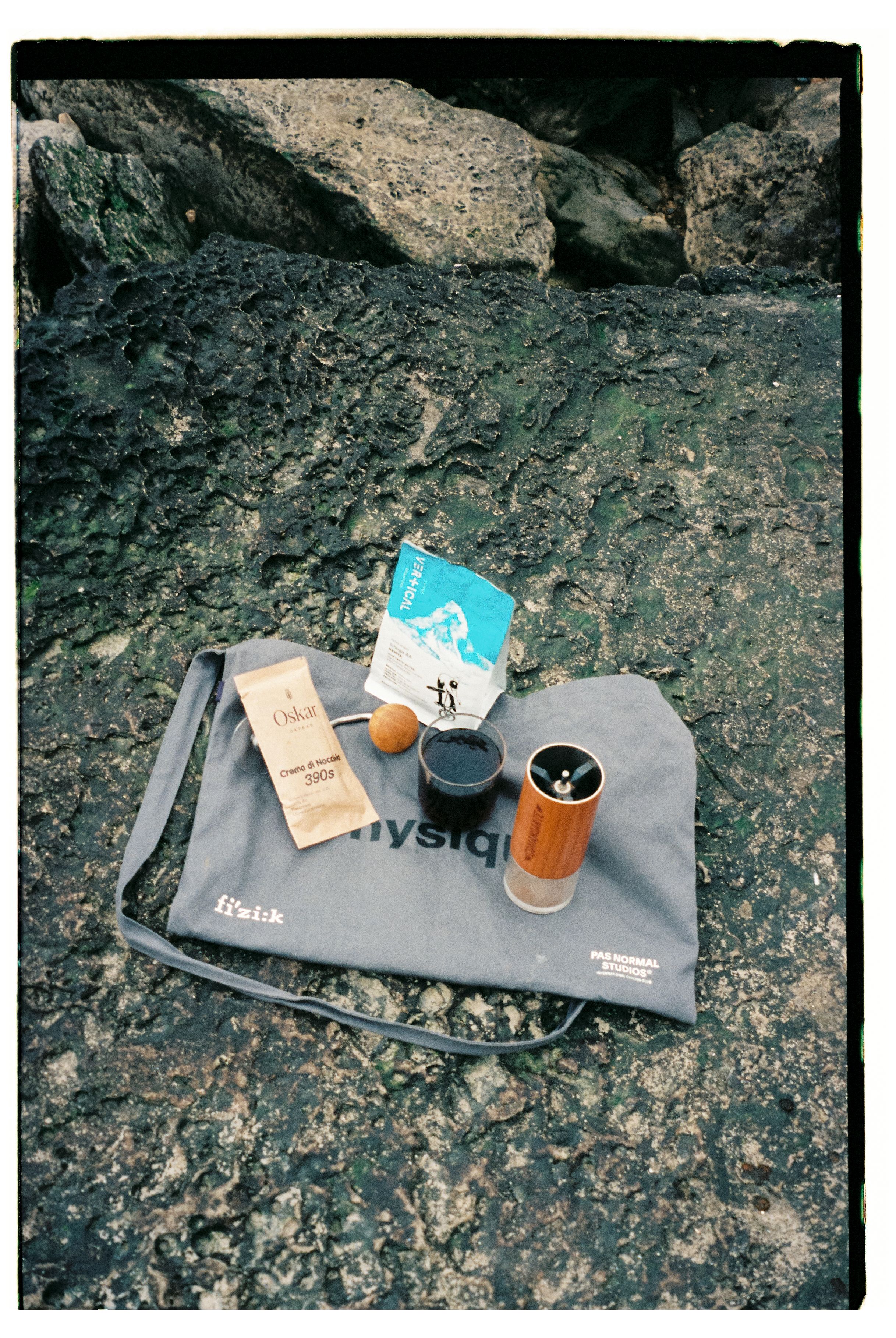 Grey Musette bag with coffee items and branding on rocky surface