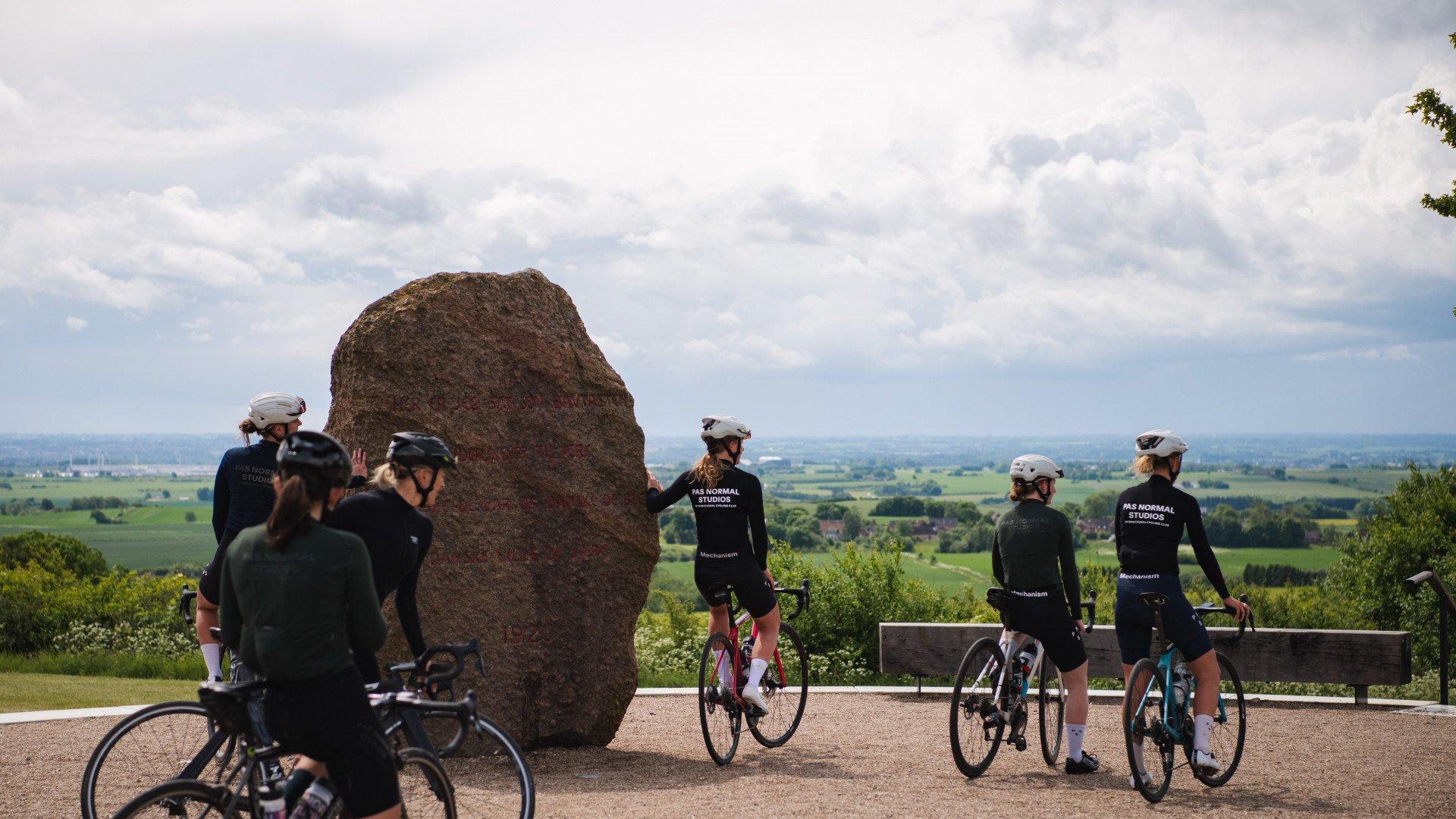 Cyclists wearing black jerseys with back lettering gathered around large stone monument with countryside vista