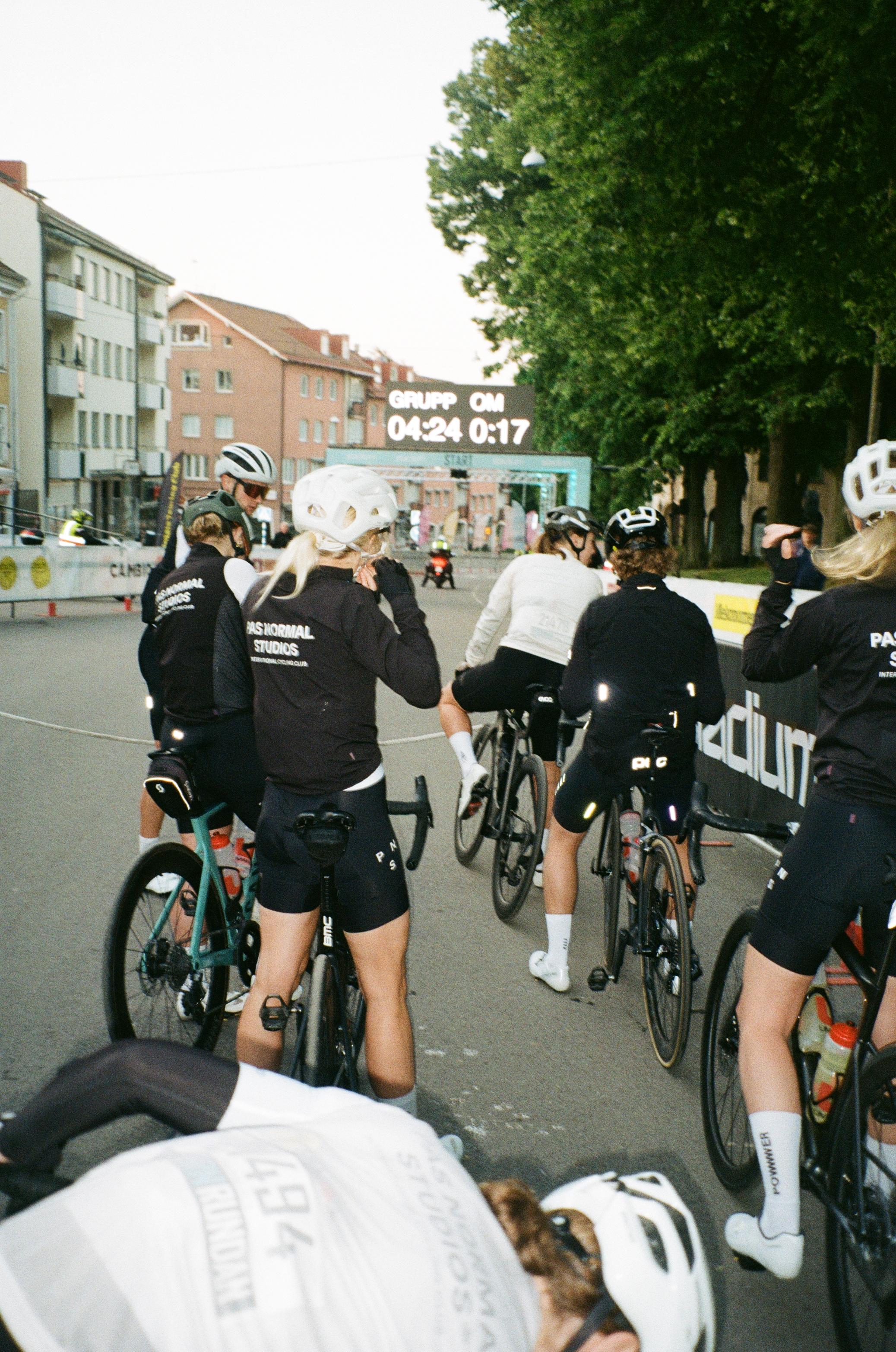 Cyclists in black jerseys and shorts at race start line with timing display showing 04:24 0:17