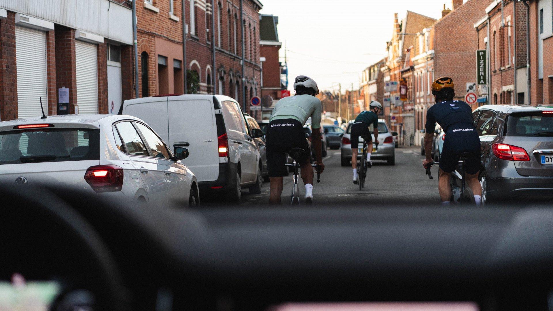Cyclists wearing Mechanism collection apparel riding through urban street with brick buildings and parked cars