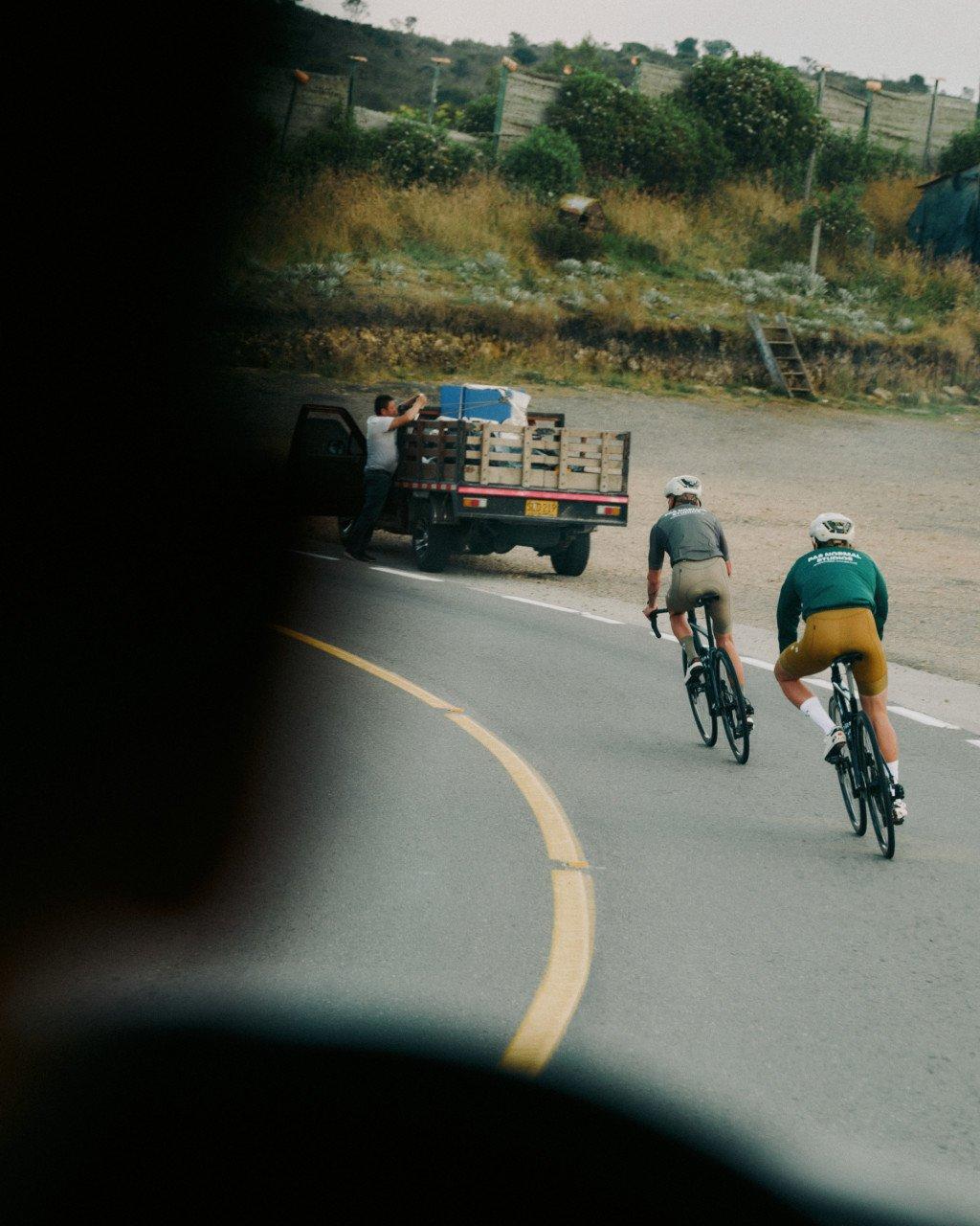 Two cyclists in green and yellow jerseys riding on mountain road with truck ahead