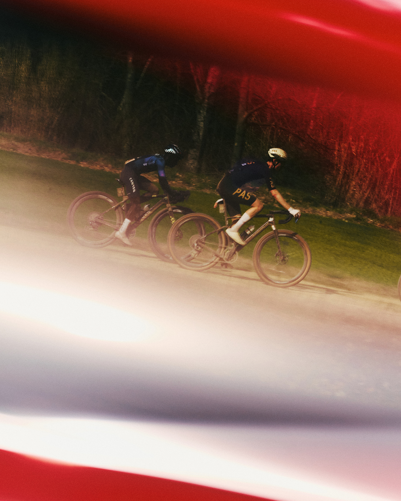 Two cyclists in dark PAS Racing jerseys riding on road with red and yellow lighting effects