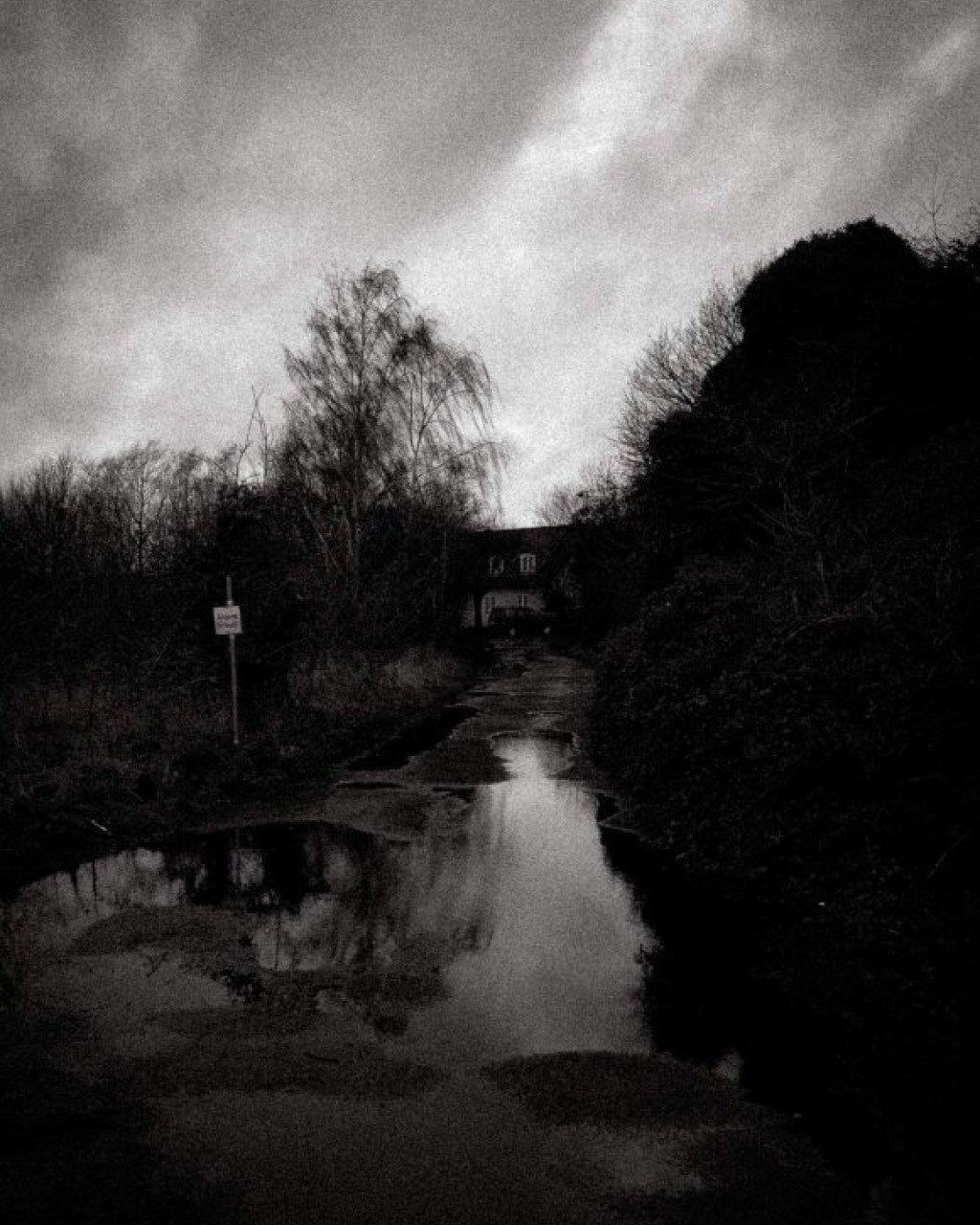 Black and white landscape showing flooded countryside path with bare trees and overcast sky