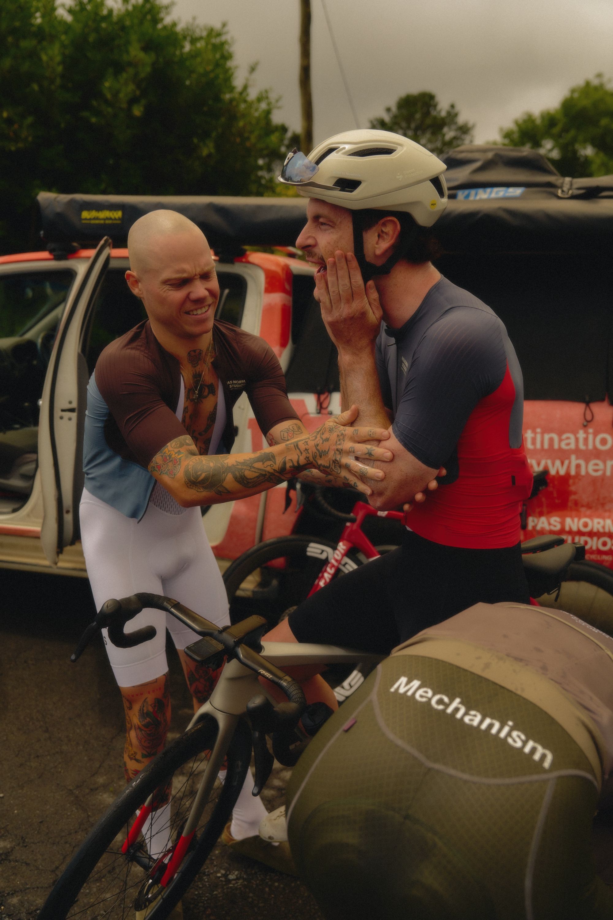 Two cyclists at race event, one in Mechanism skinsuit, one in dark jersey with red details