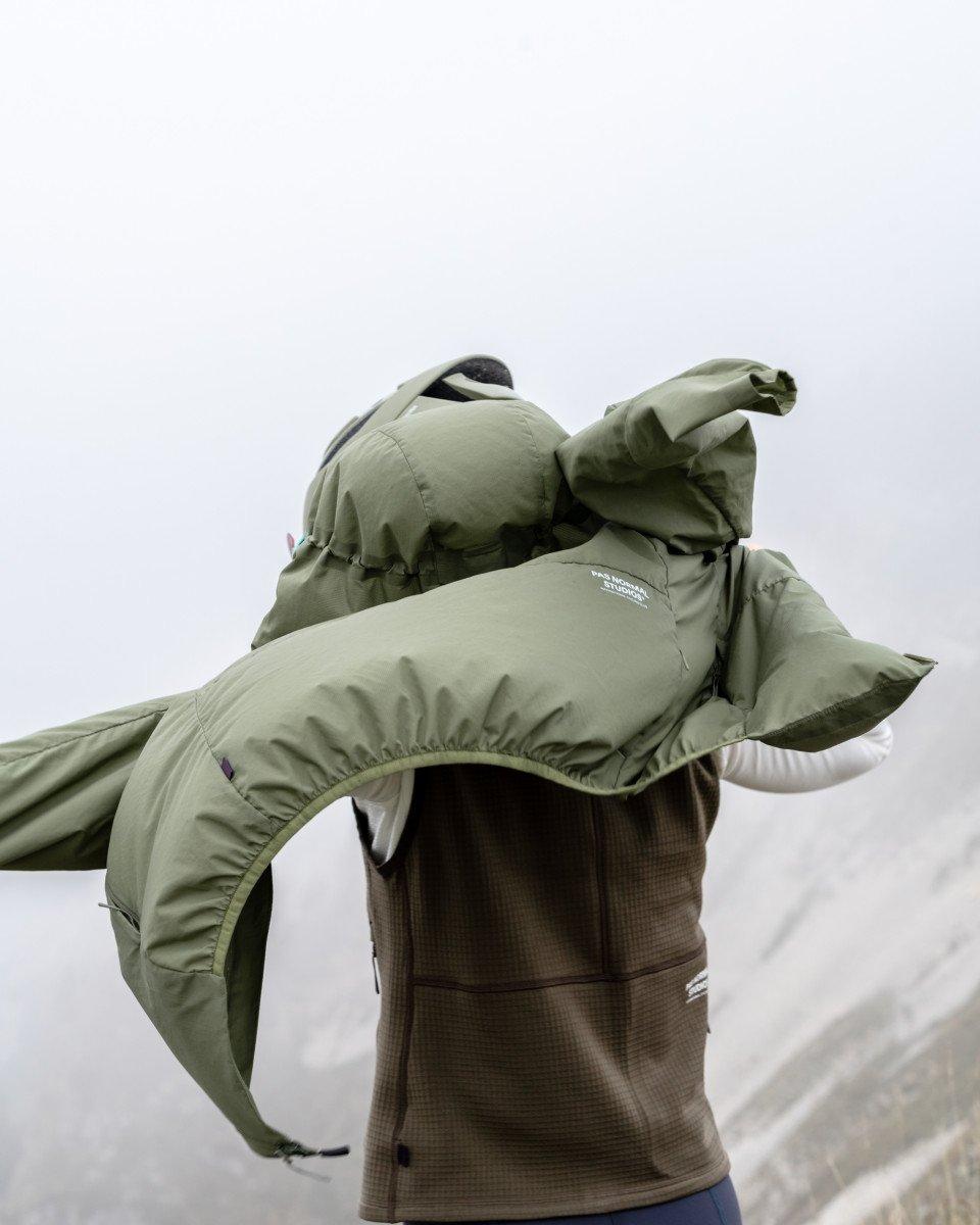 Model wearing green insulated jacket over brown base layer in mountain setting